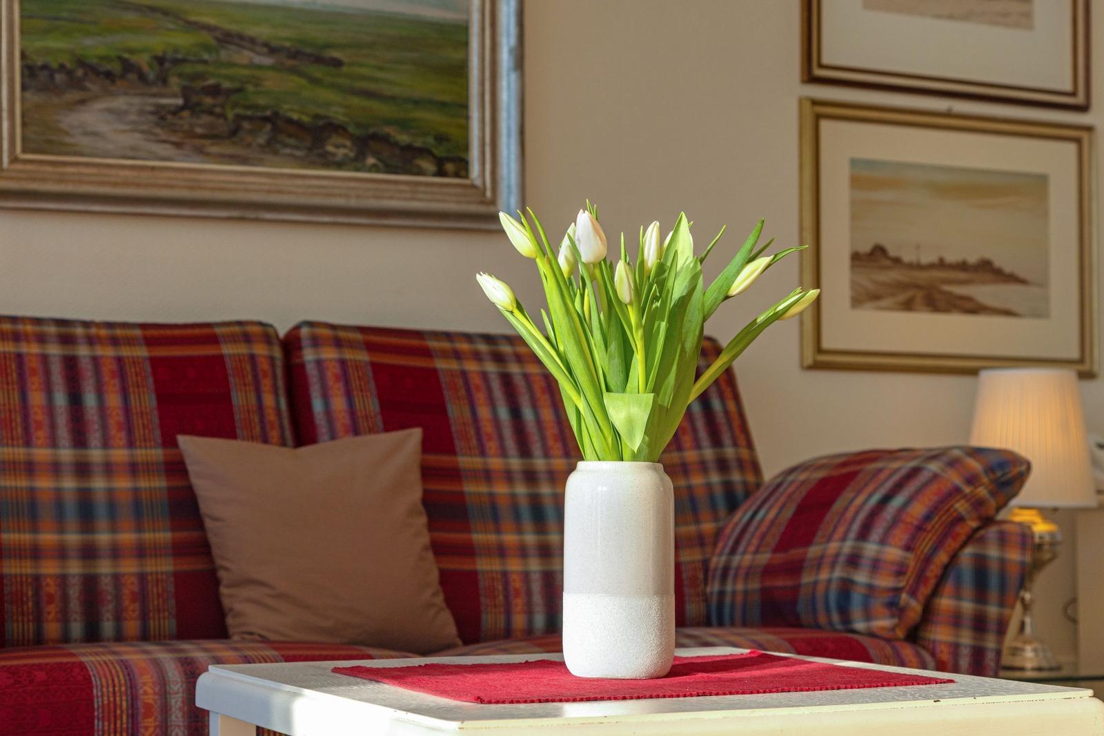 Ein weißer Vasenstrauß Tulpen steht auf einem Tisch vor einem Sofa mit kariertem Bezug.