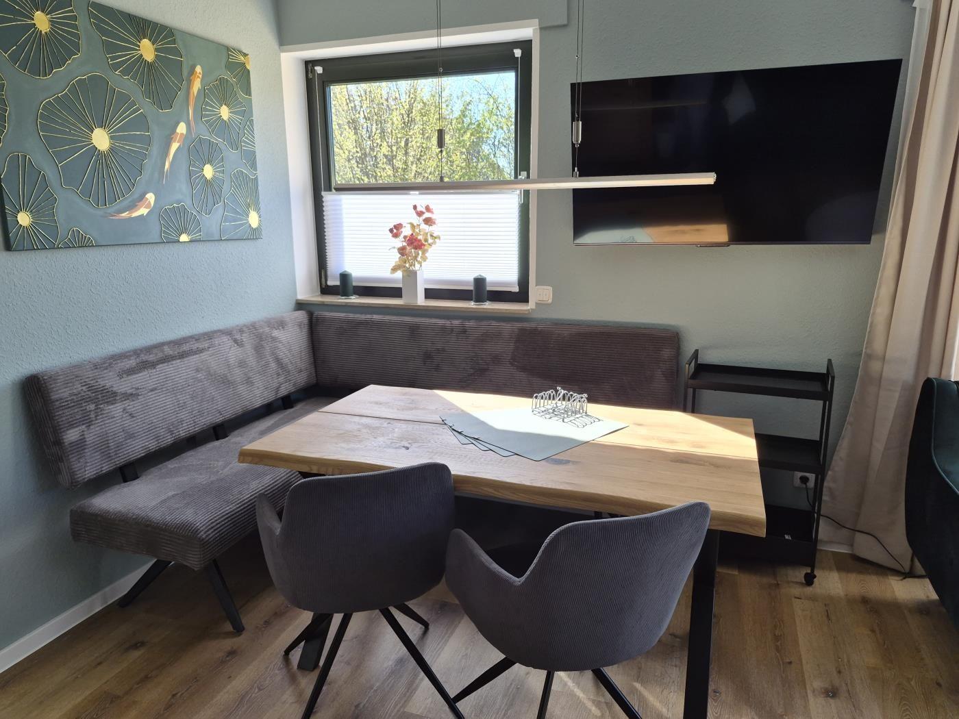 Essbereich mit Holztisch, Sitzbank und zwei Stühlen. Fenster und Wandbild sichtbar.