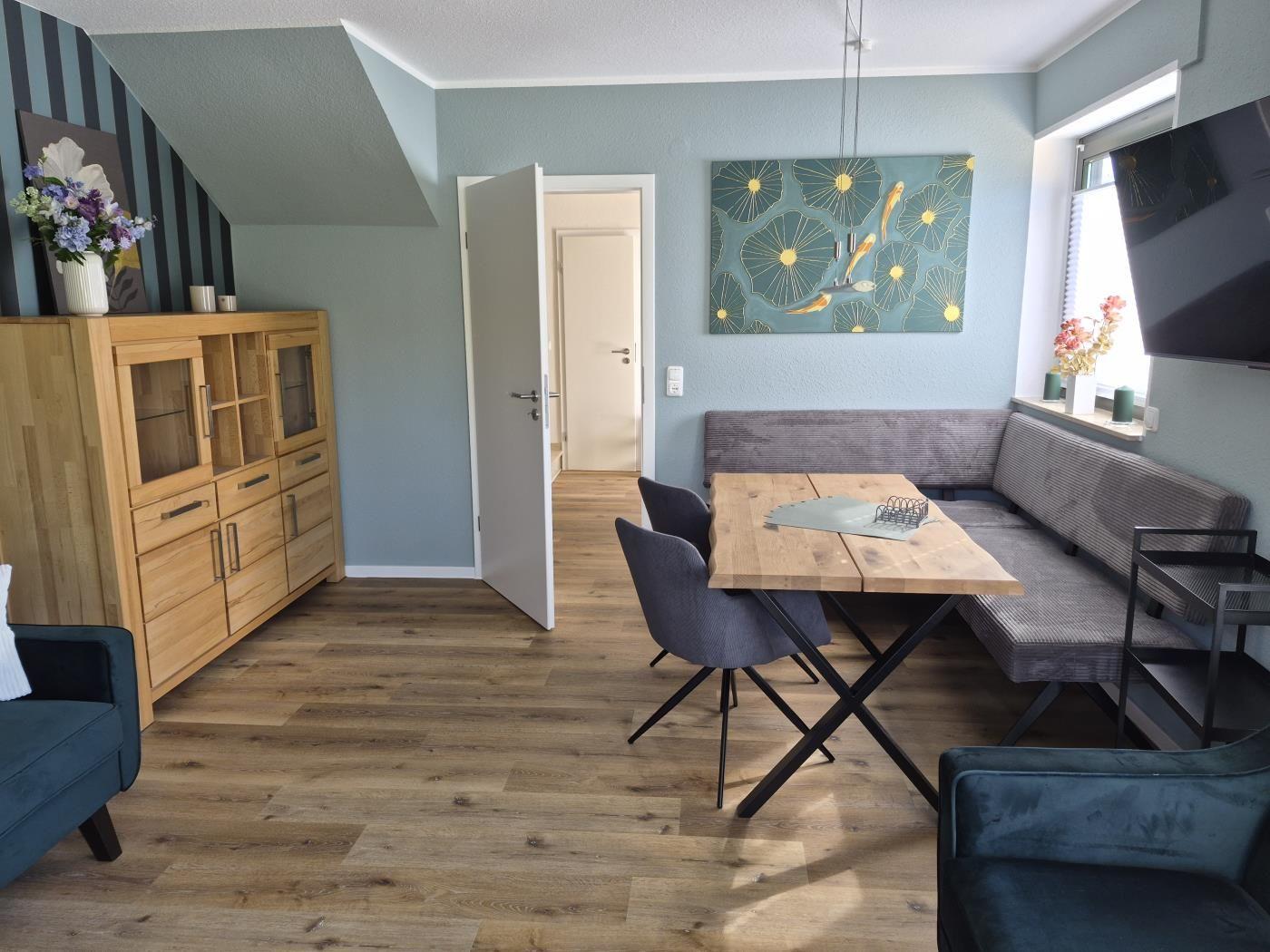 Essbereich mit Holztisch, grauen Stühlen und Sofa. Wandbild und TV sichtbar.
