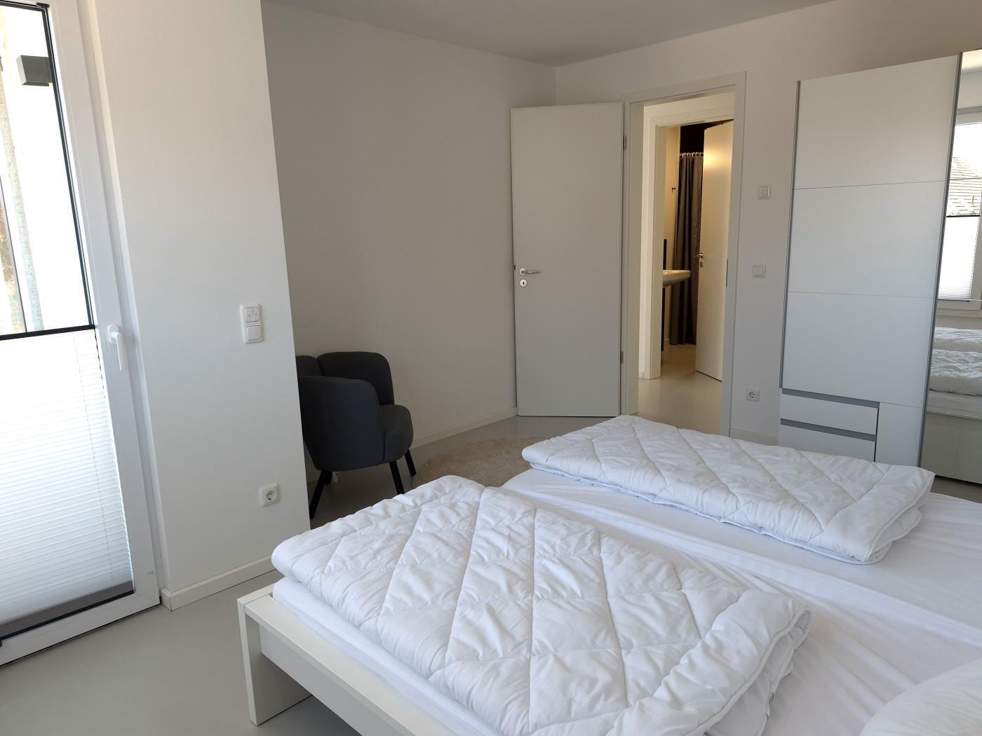 Schlafzimmer mit weißem Bett, grauer Sessel und Schrank. Tür führt in Badezimmer.