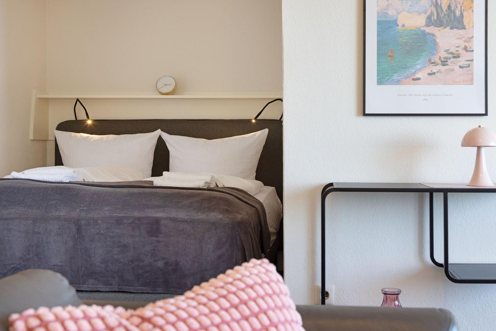 Schlafzimmer mit Bett, Kissen, Wandbild und Tisch mit Lampe.