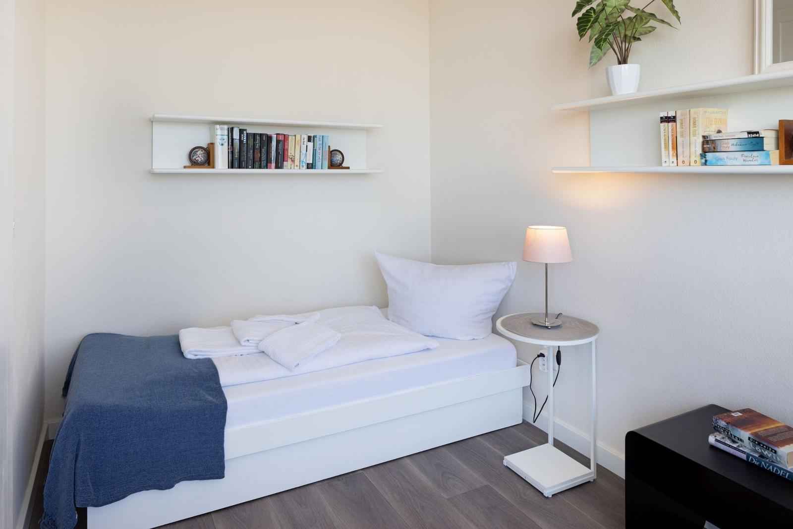 Schlafzimmer mit Bett, Nachttisch, Lampe und Regalen mit Büchern und Pflanze.