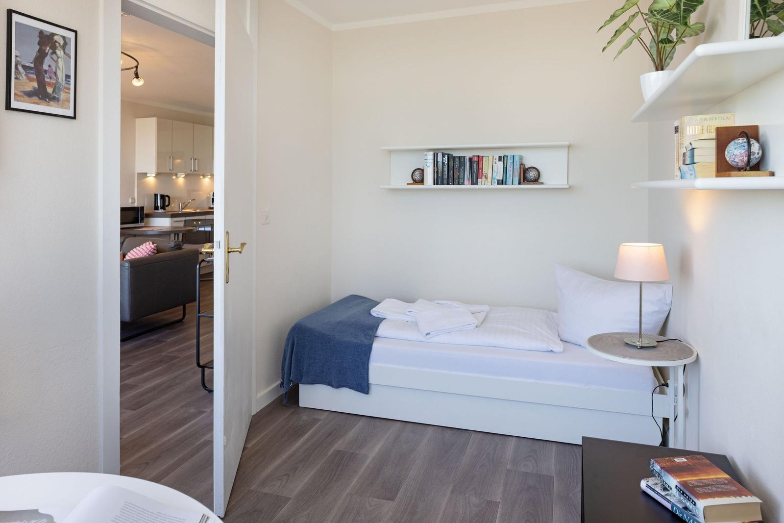 Schlafzimmer mit Bett, Nachttisch und Regal. Durchgang zur Küche sichtbar.