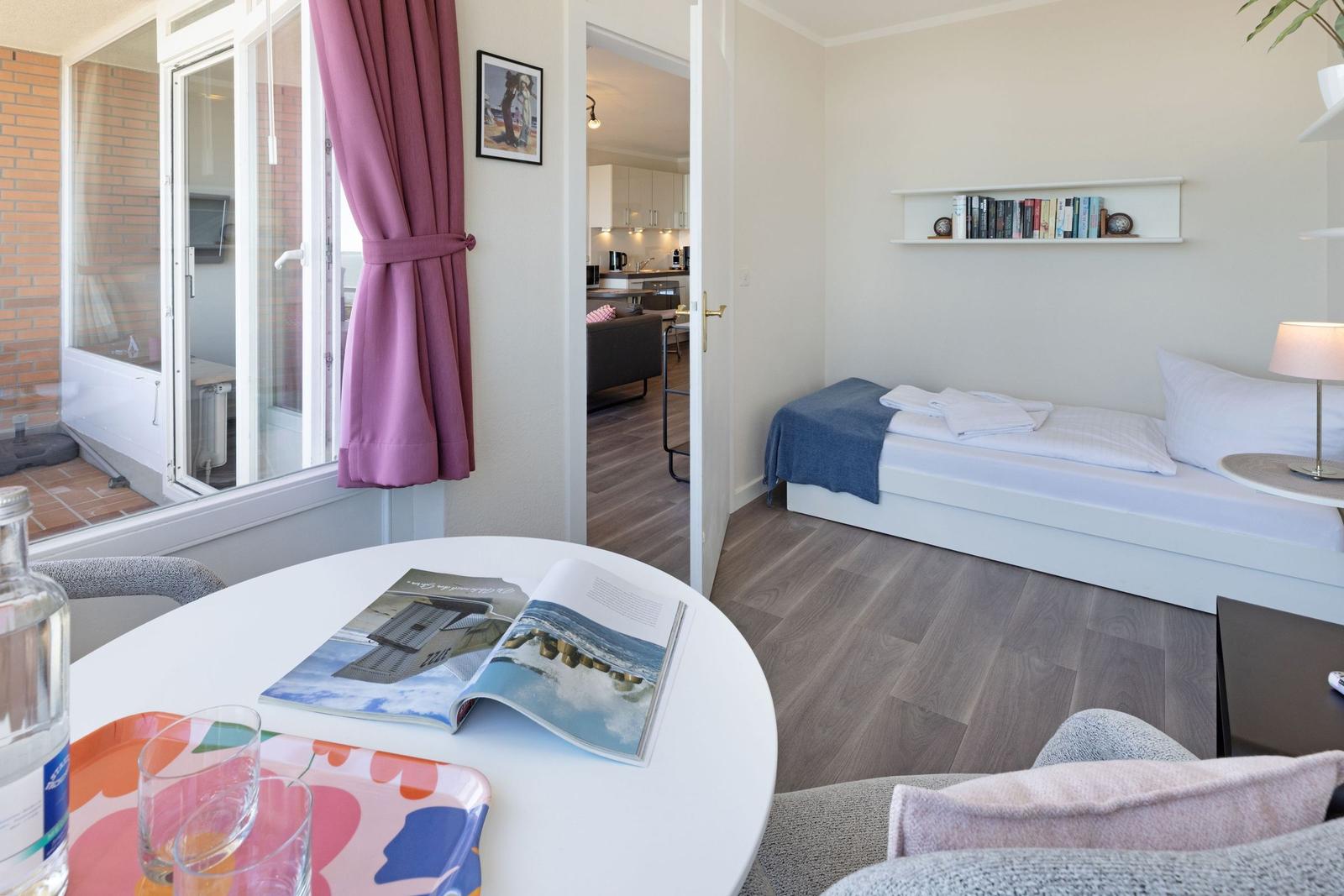 Ein Schlafzimmer mit Bett, Nachttisch und Bücherregal. Durchgang zur Küche und Terrasse mit Vorhang.