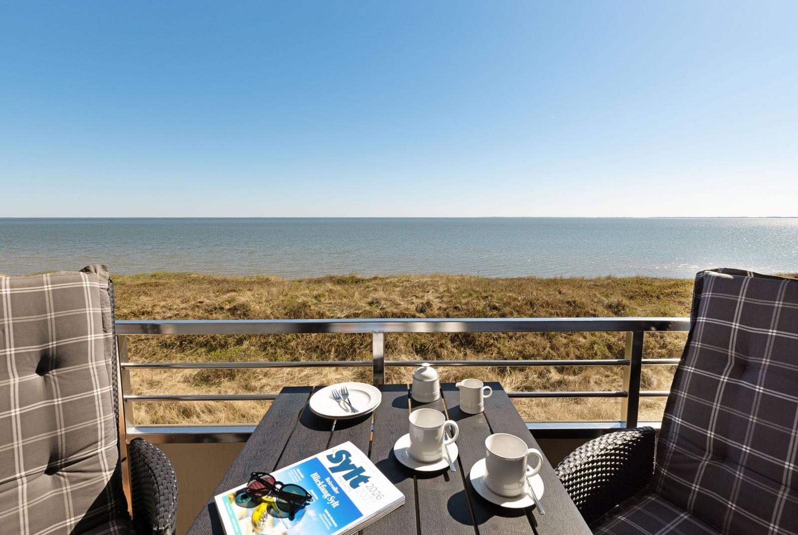 Terrasse mit Tisch, Stühlen und Meerblick. Auf dem Tisch stehen Tassen und eine Zeitschrift.