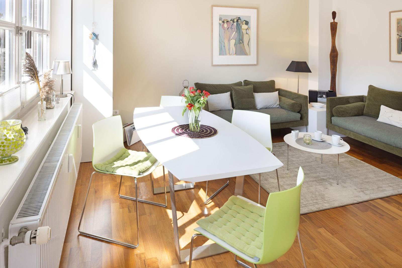 Dining area with white table and green chairs. Living area with sofa and couch. Wooden floor and windows.