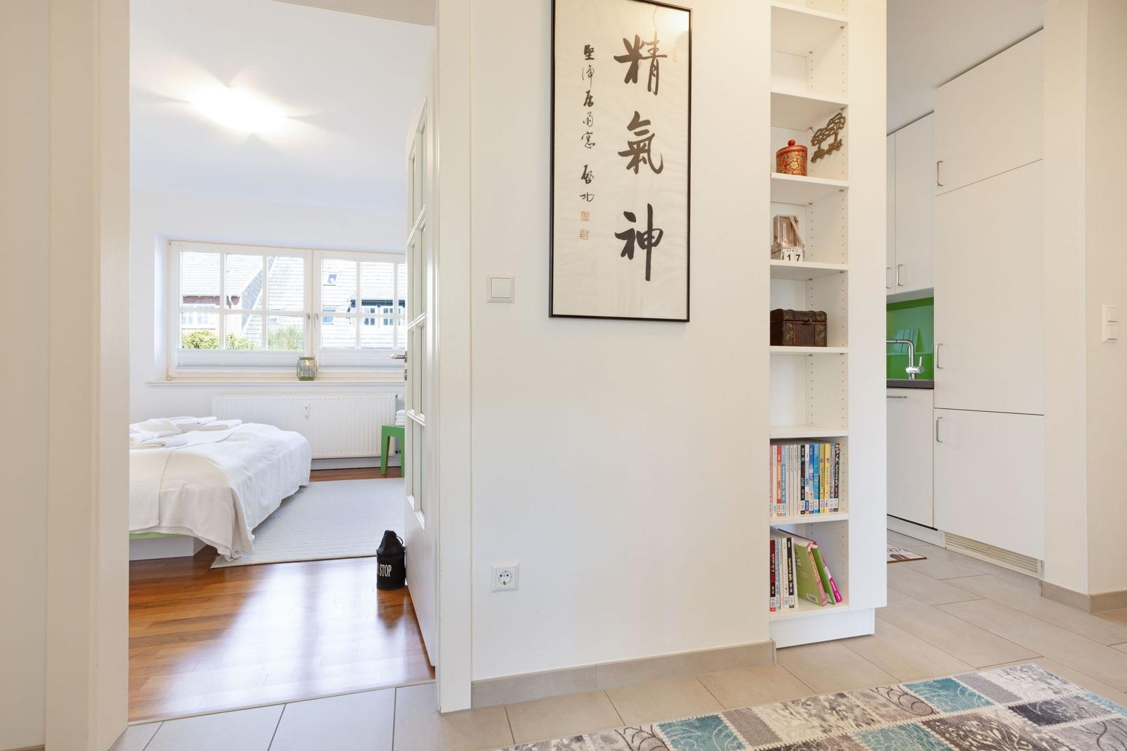 Bedroom with bed and window. Kitchen with white cabinets. Wall with calligraphy and bookshelf.