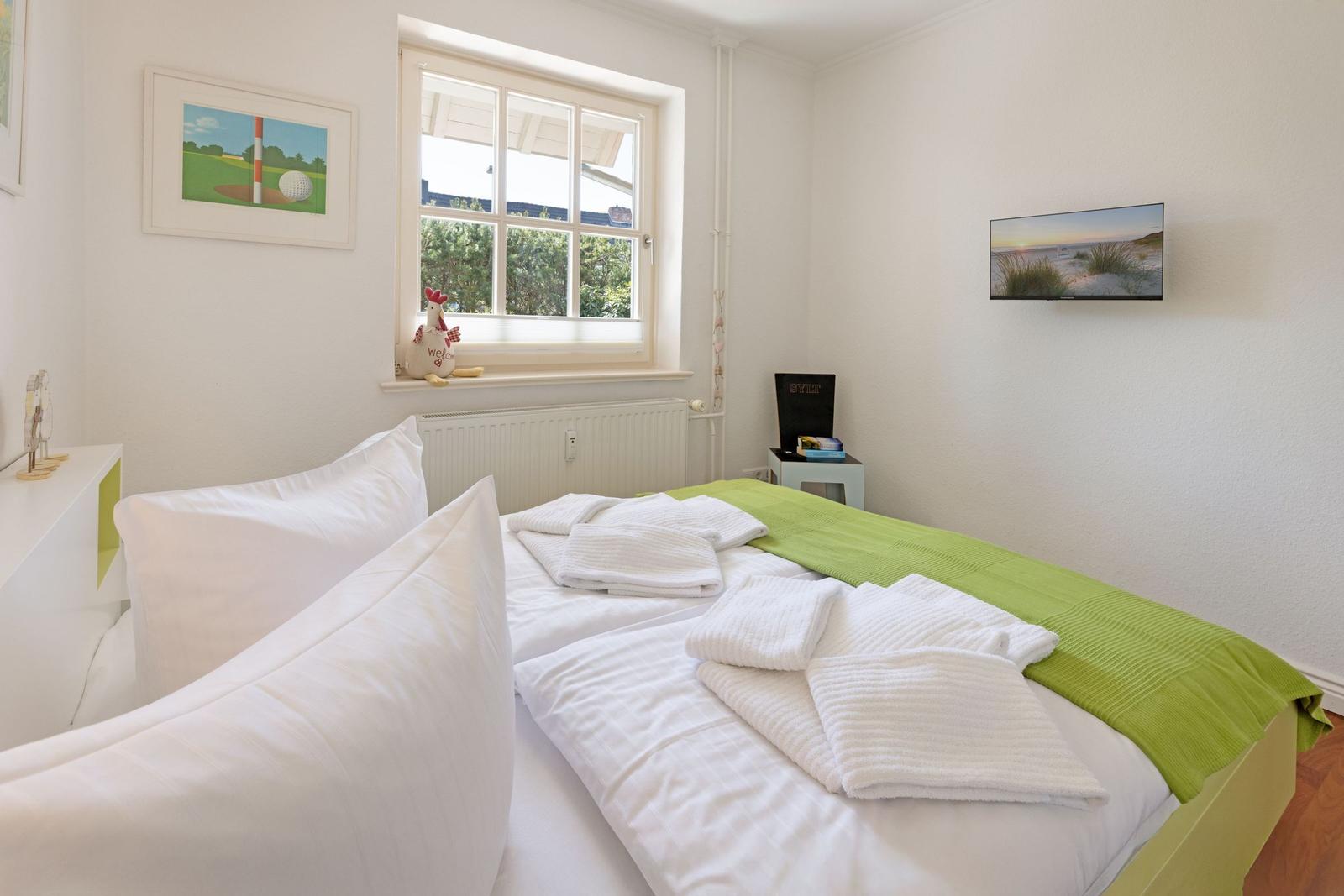 A bedroom with white bedding, green blanket edge, and view of greenery through the window.