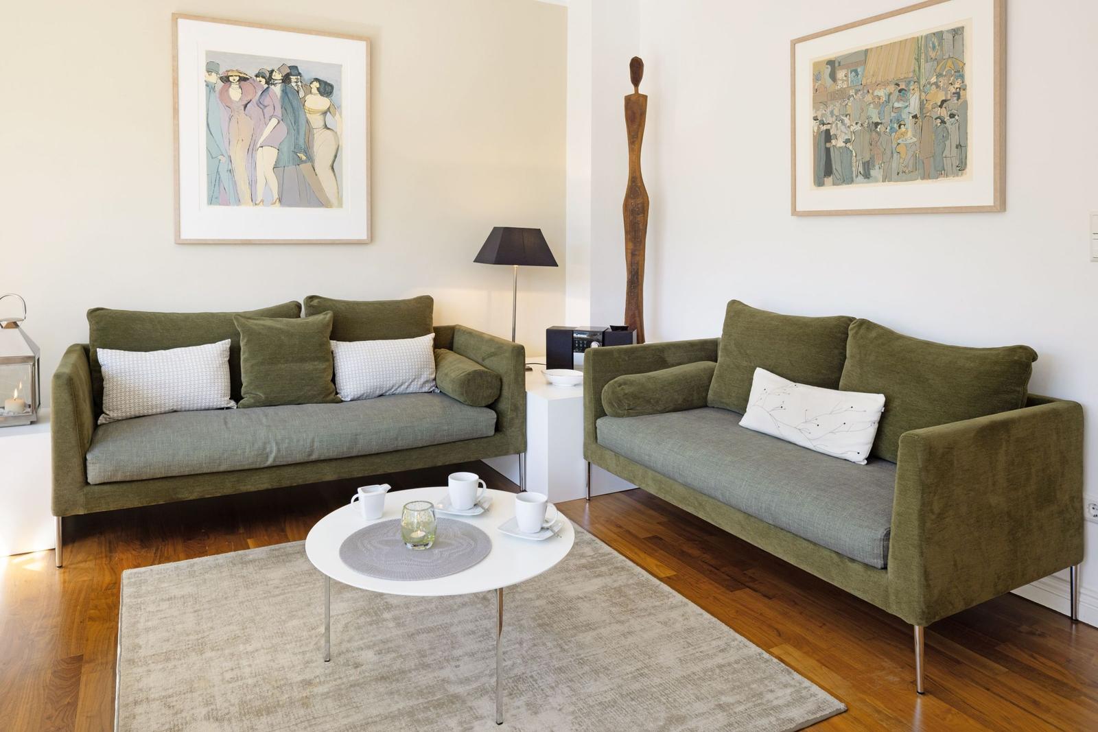 Two green sofas with cushions, round table and wall art in a living room with hardwood floor.