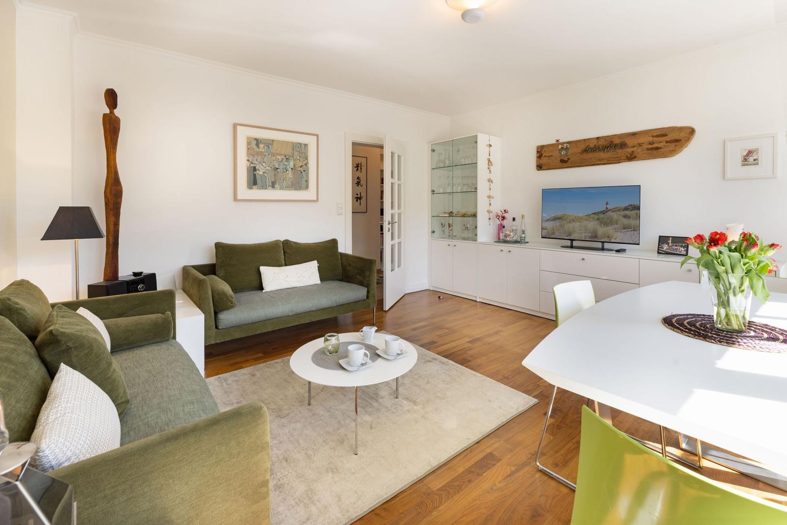 Living and dining area with green sofas, wooden floor, and TV. Table with flowers and side table.