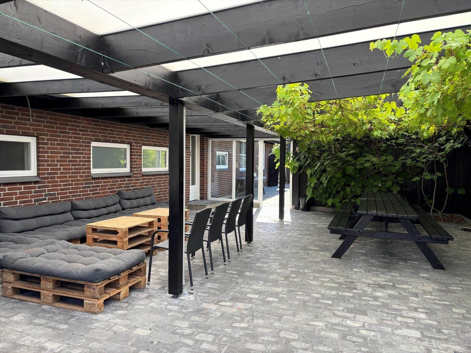 Terrace with seating and table under a pavilion. Background: brick wall and plants.