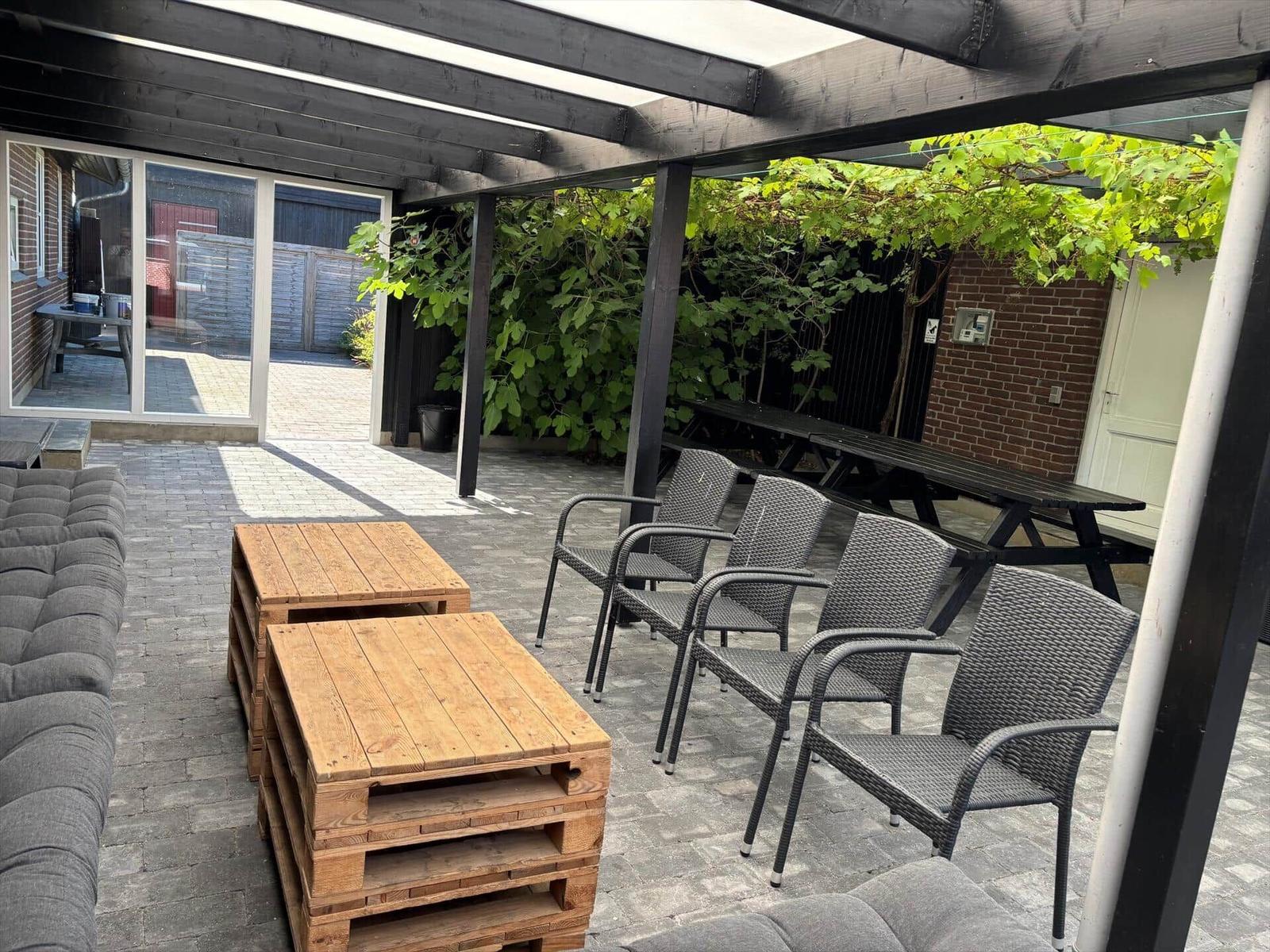 Terrace with seating, wooden tables, and pergola. Greenery and buildings in the background.