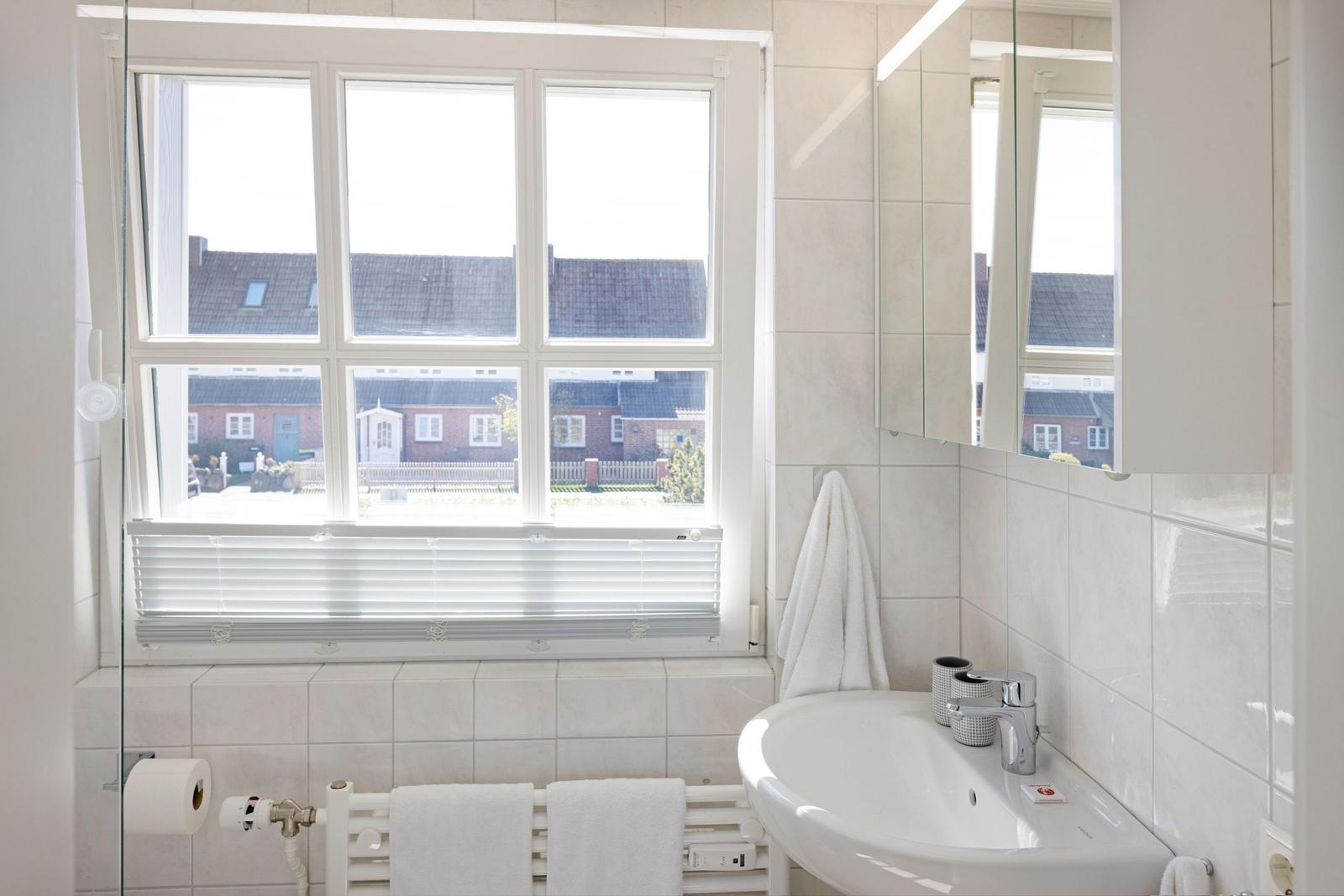 Badezimmer mit Fenster, Spiegel und Waschbecken. Weiße Fliesen und Handtücher.