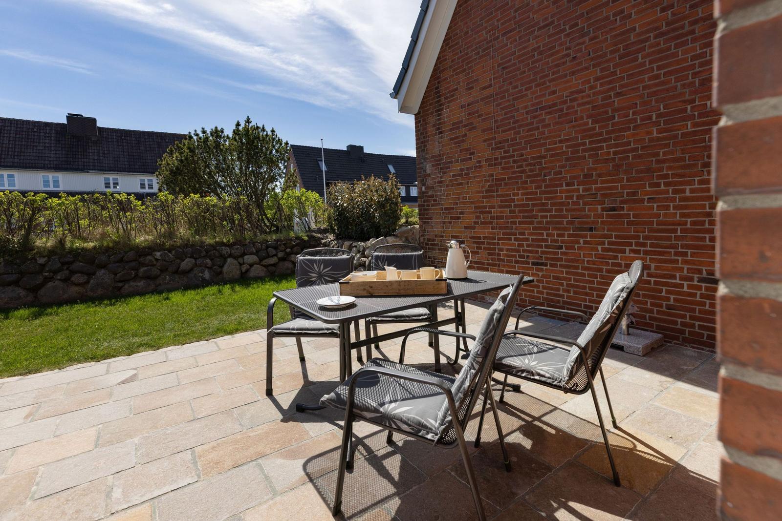 Terrasse mit Tisch und Stühlen neben rotem Backsteinhaus. Grünflächen und Steinmauer im Hintergrund.