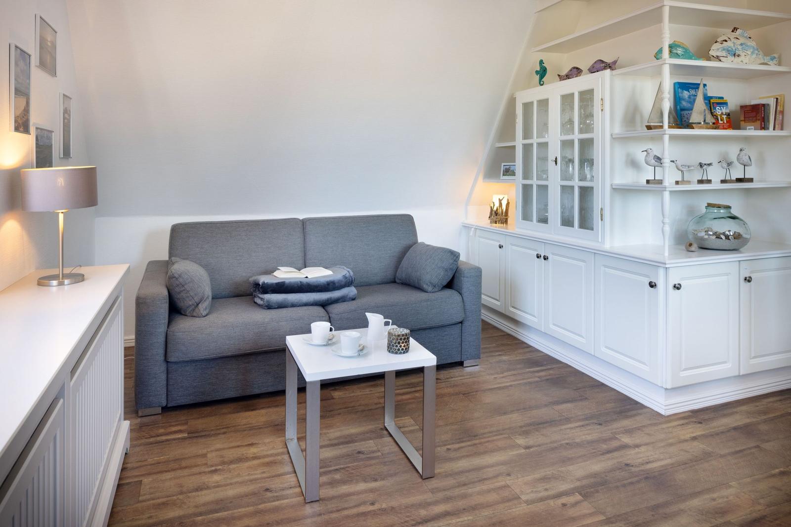 Ein Wohnzimmer mit grauem Sofa, weißem Tisch und weißen Schränken. Holzfußboden und Wandregale.