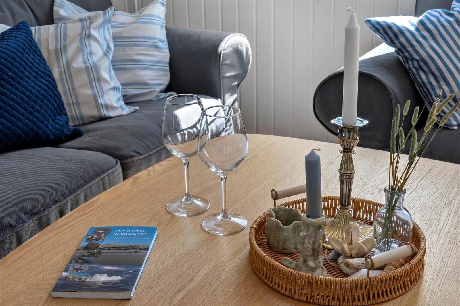 Holztisch mit Weinflaschen, Kerzen und Buch vor einem Sofa mit Kissen.