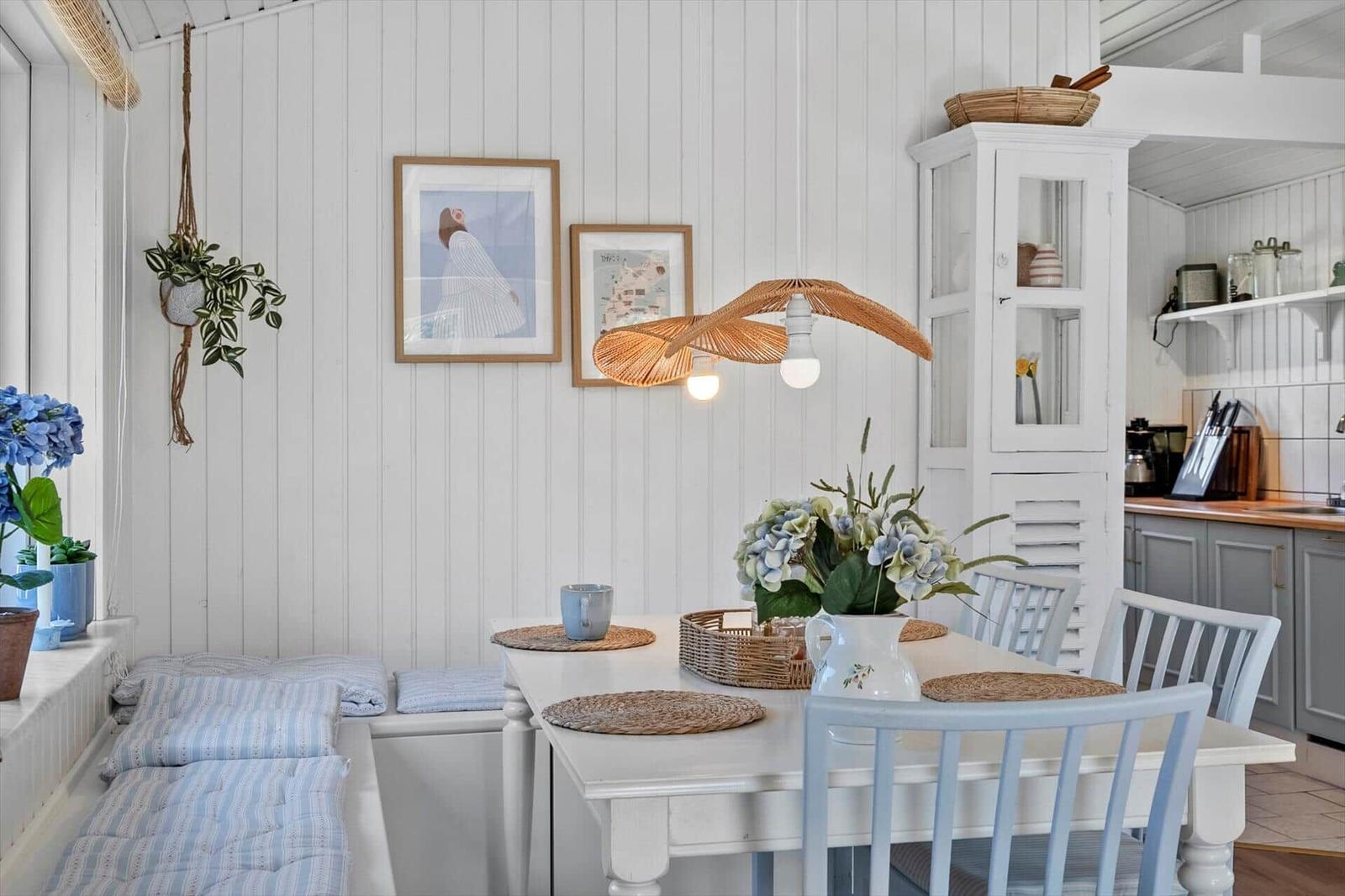 Essbereich mit weißem Tisch, blauen Stühlen und Blumenarrangement. Hintergrund: weiße Wände und Küchenbereich.
