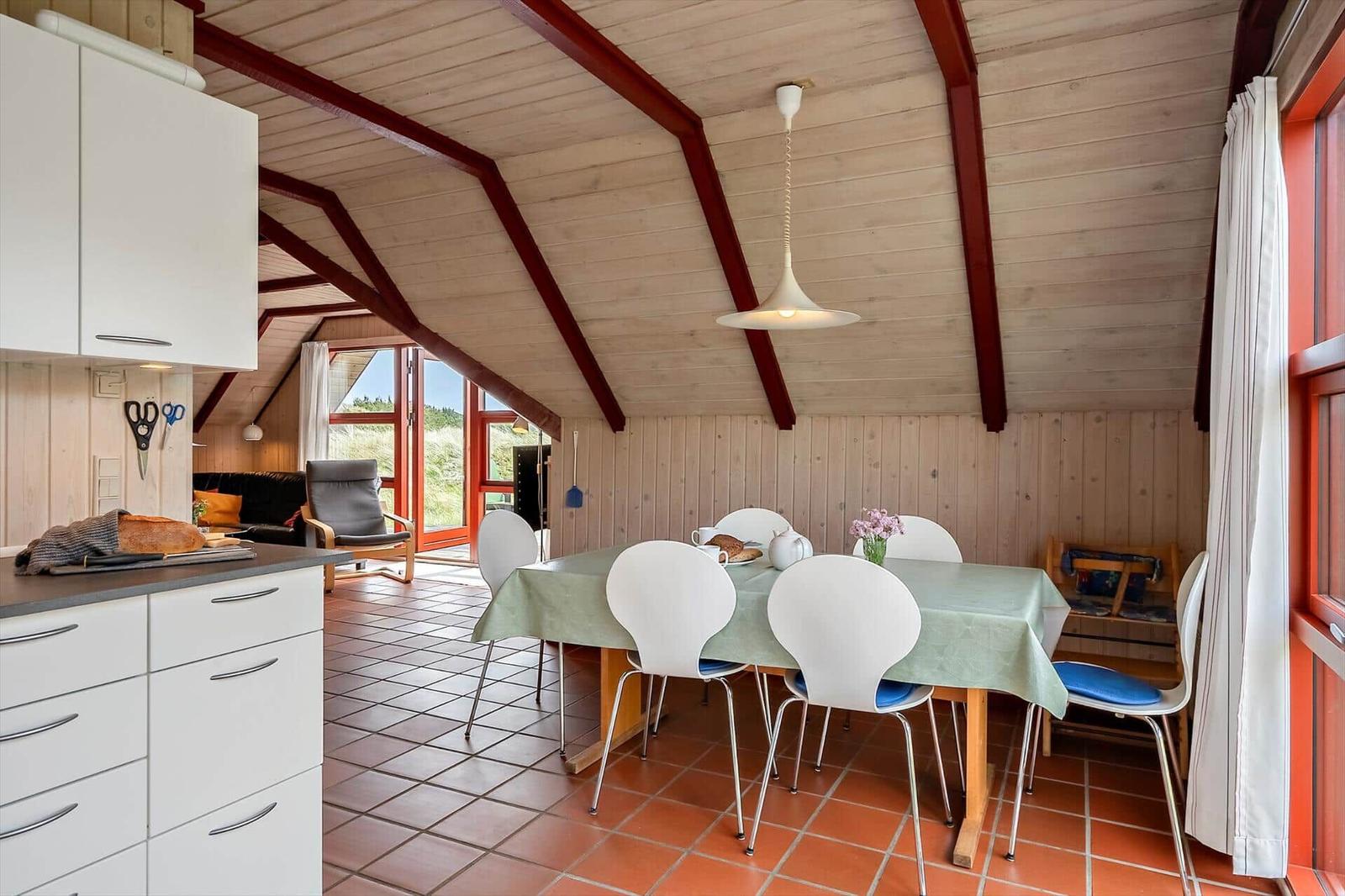 Essbereich mit Tisch, Stühlen und Küchenzeile unter Holzdecke mit roten Trägern.