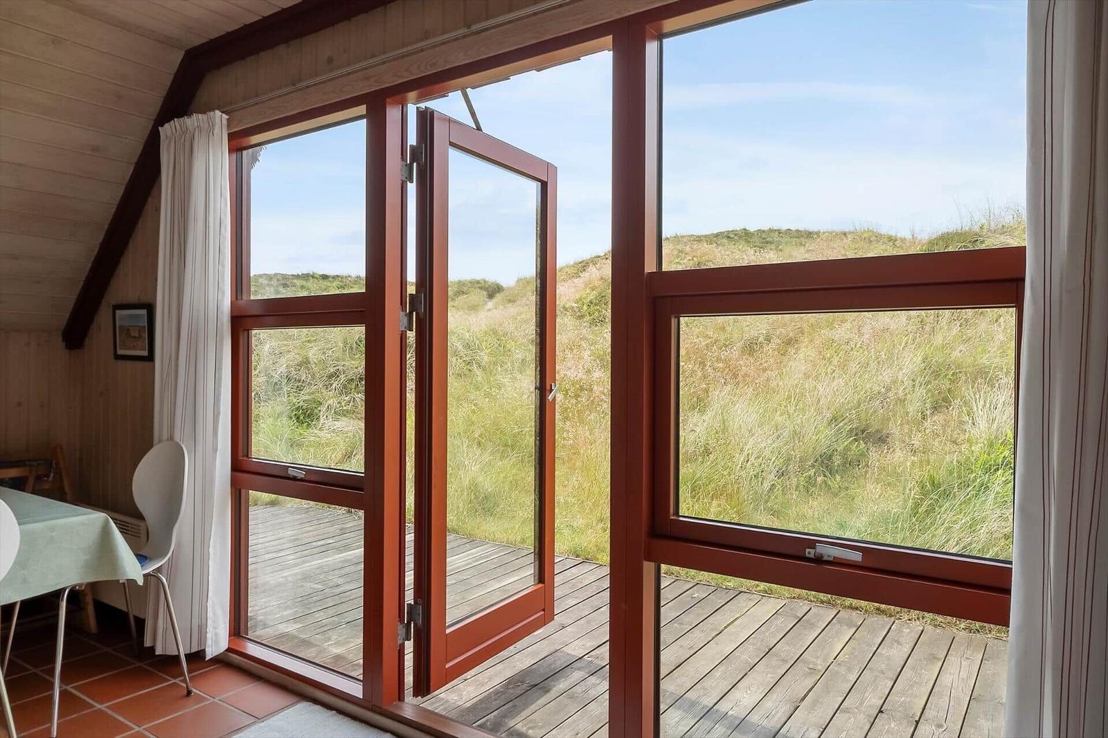 Durch große Fenster mit rotem Rahmen blickt man auf eine Graslandschaft. Im Innenraum steht ein Tisch mit Stühlen.