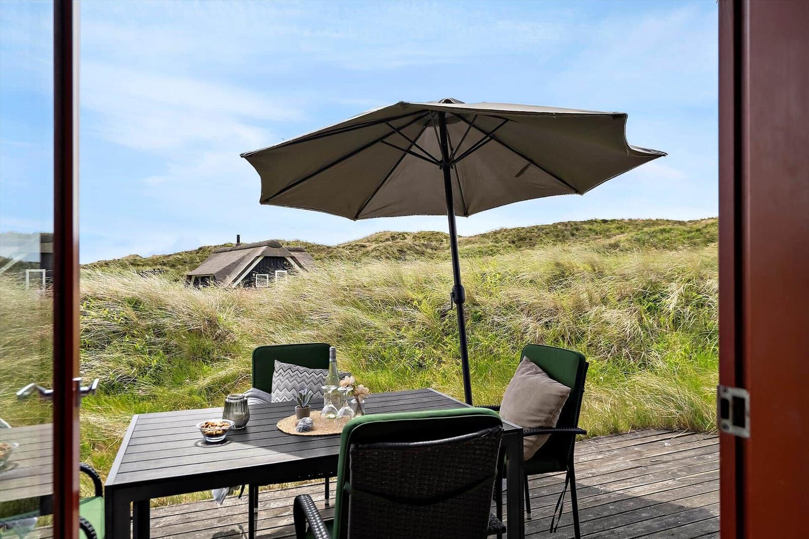 Terrasse mit Tisch, Stühlen und Sonnenschirm. Blick auf Grasland und Ferienhaus.