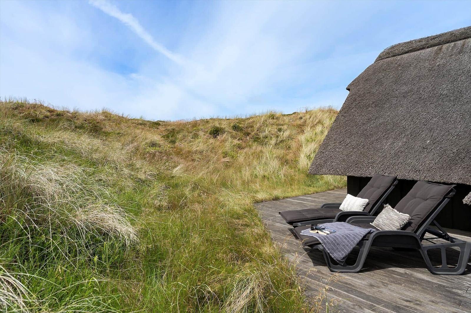 Zwei Liegestühle stehen auf einer Holterrasse neben einem strohgedeckten Haus. Grasbewachsene Hügel im Hintergrund.