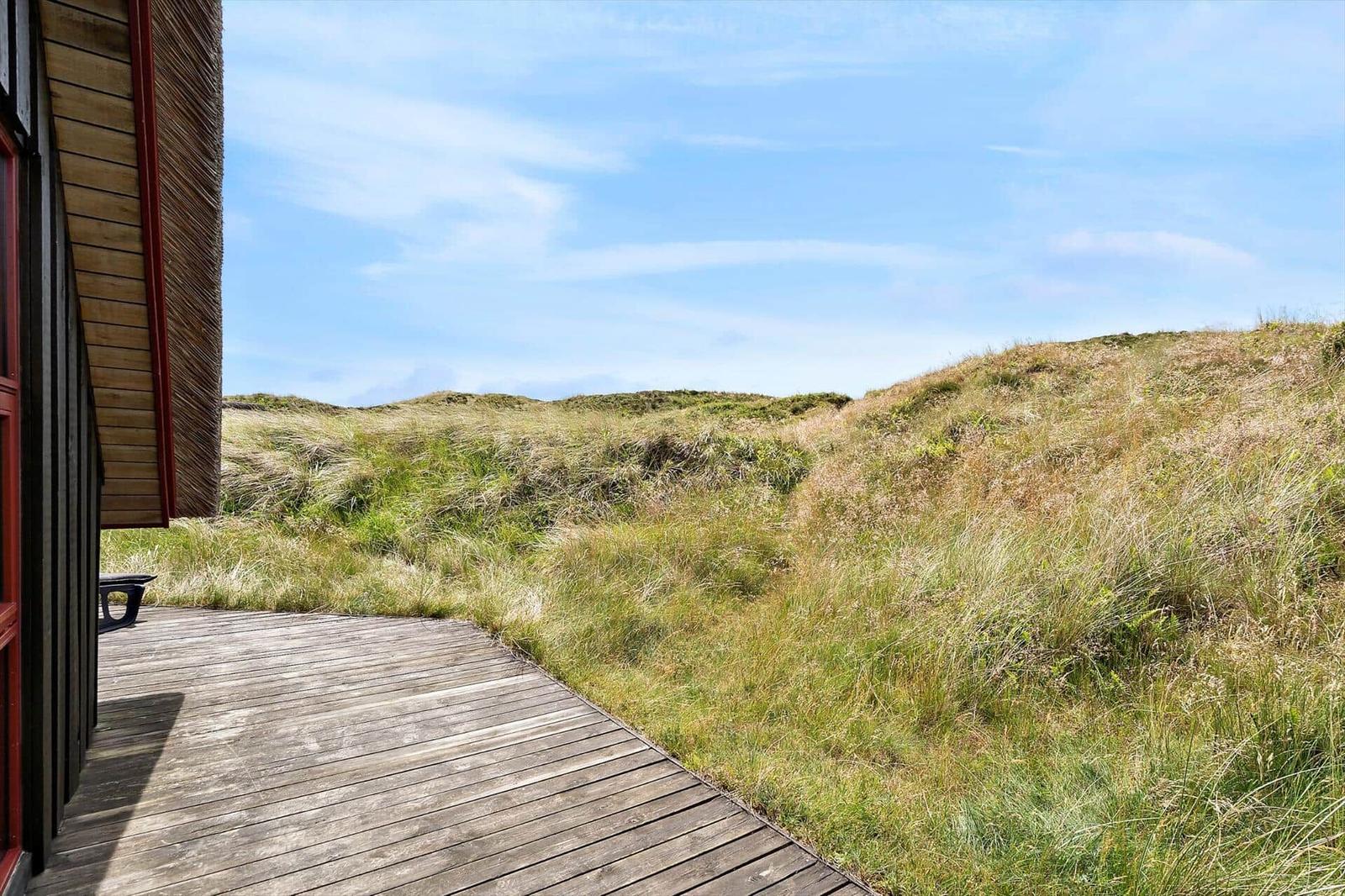 Holzterrasse führt zu grasbewachsenen Dünen unter blauem Himmel.