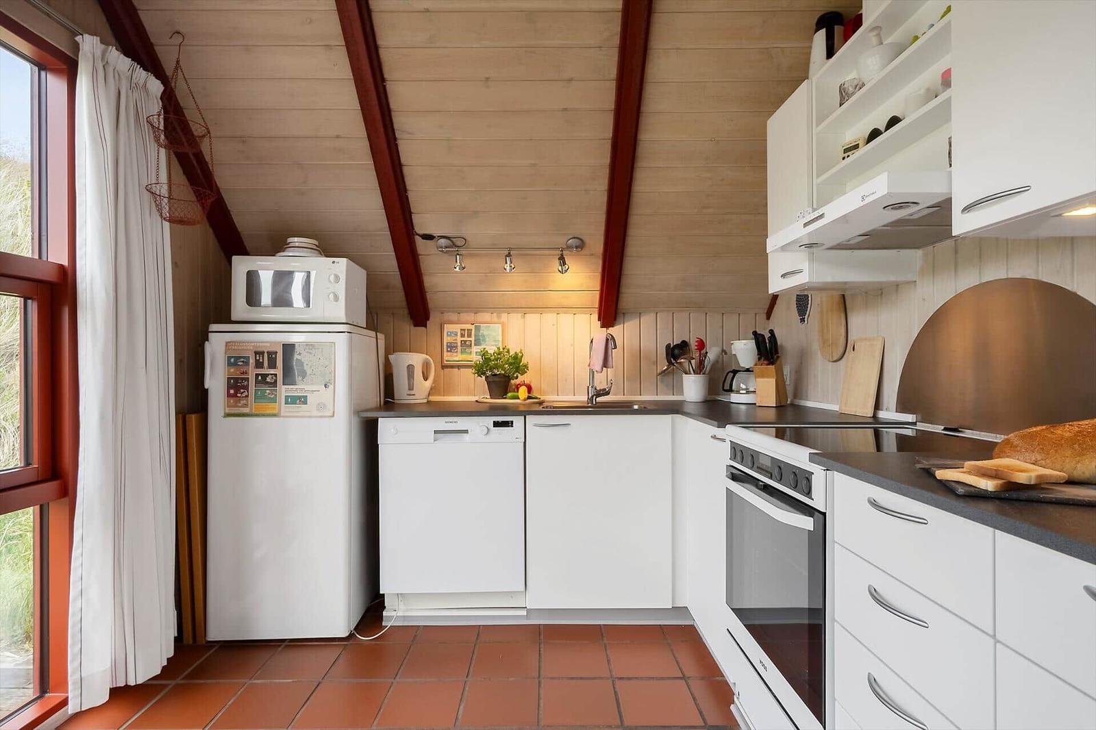 Küche mit weißen Schränken, Kühlschrank, Spüle und Herd. Holzdecke und Fenster mit Vorhang.