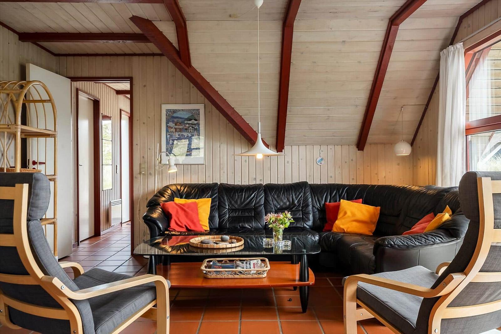 Wohnzimmer mit schwarzer Ledercouch, Holzstühlen und Glas-Tisch. Holzwände und Deckenbalken.