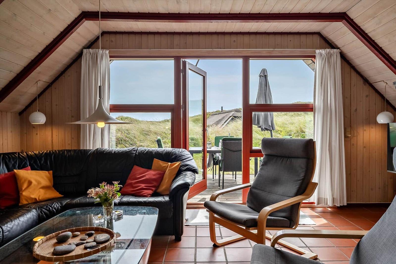 Wohnzimmer mit Sofa, Sessel und Blick auf Terrasse und Dünen.