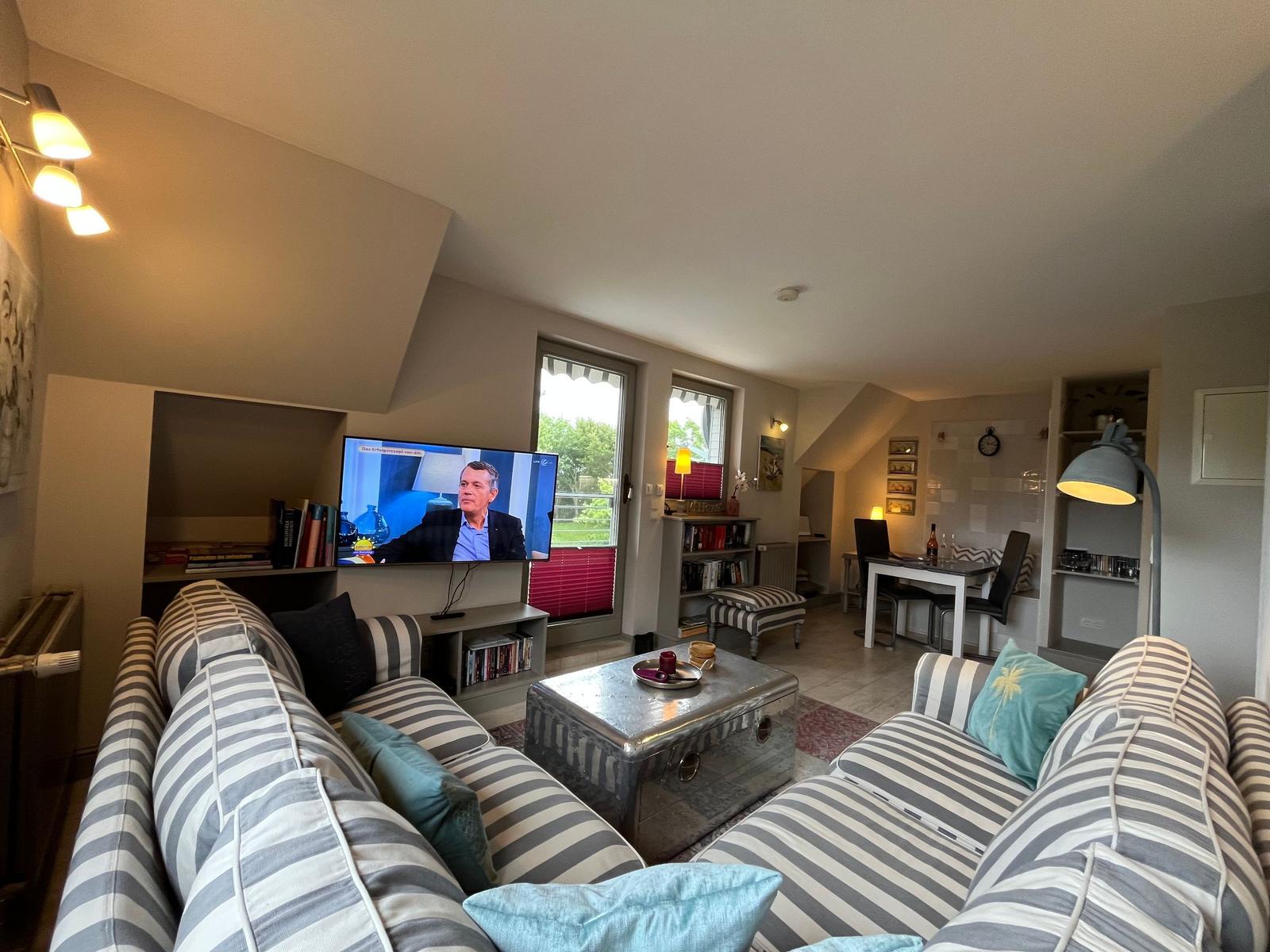 Living area with sofa, TV, and dining area. Windows with garden view.