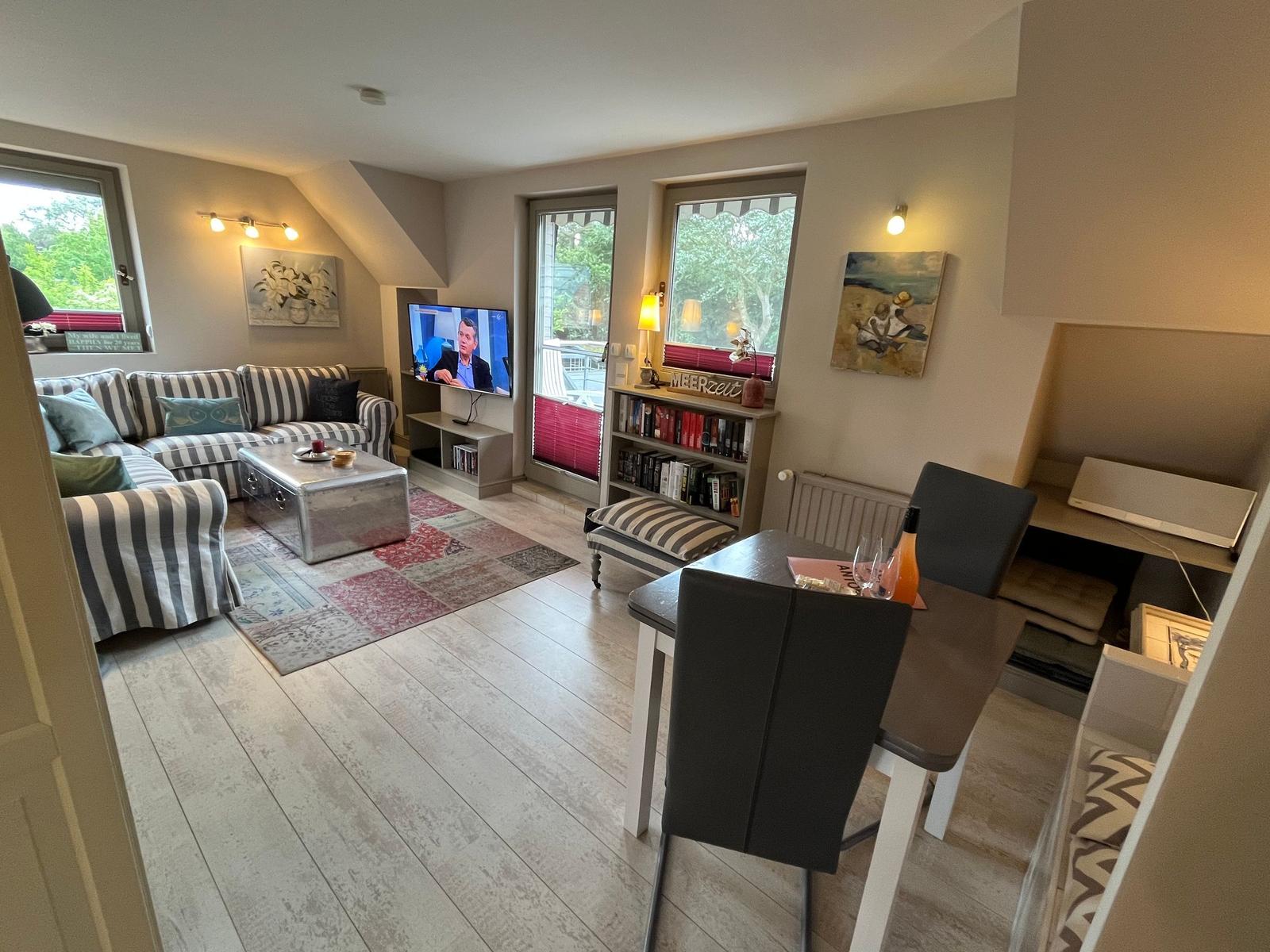 Living and dining area with sofa, TV, table and chairs. Windows with view of greenery.