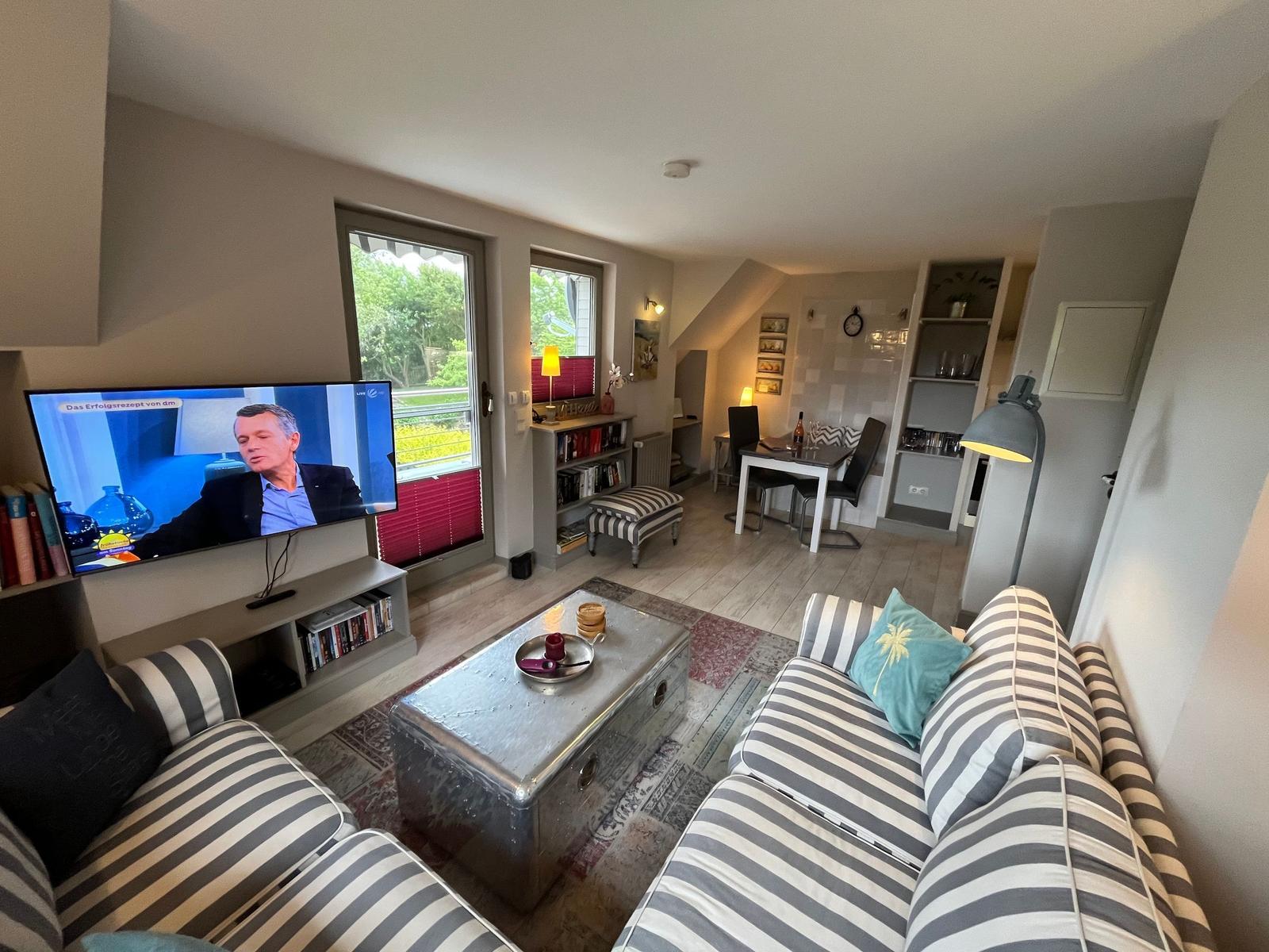 Living room with sofa, TV, and dining area. Windows overlook the garden.