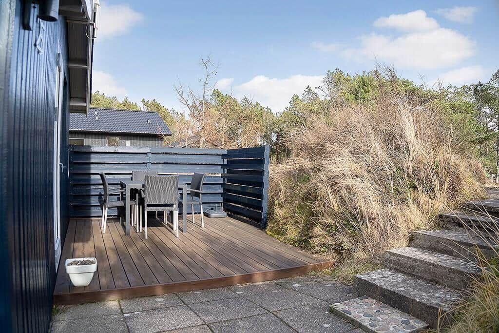 Terrasse mit Tisch und Stühlen neben einem blauen Haus. Hintergrund: Gras und Bäume.