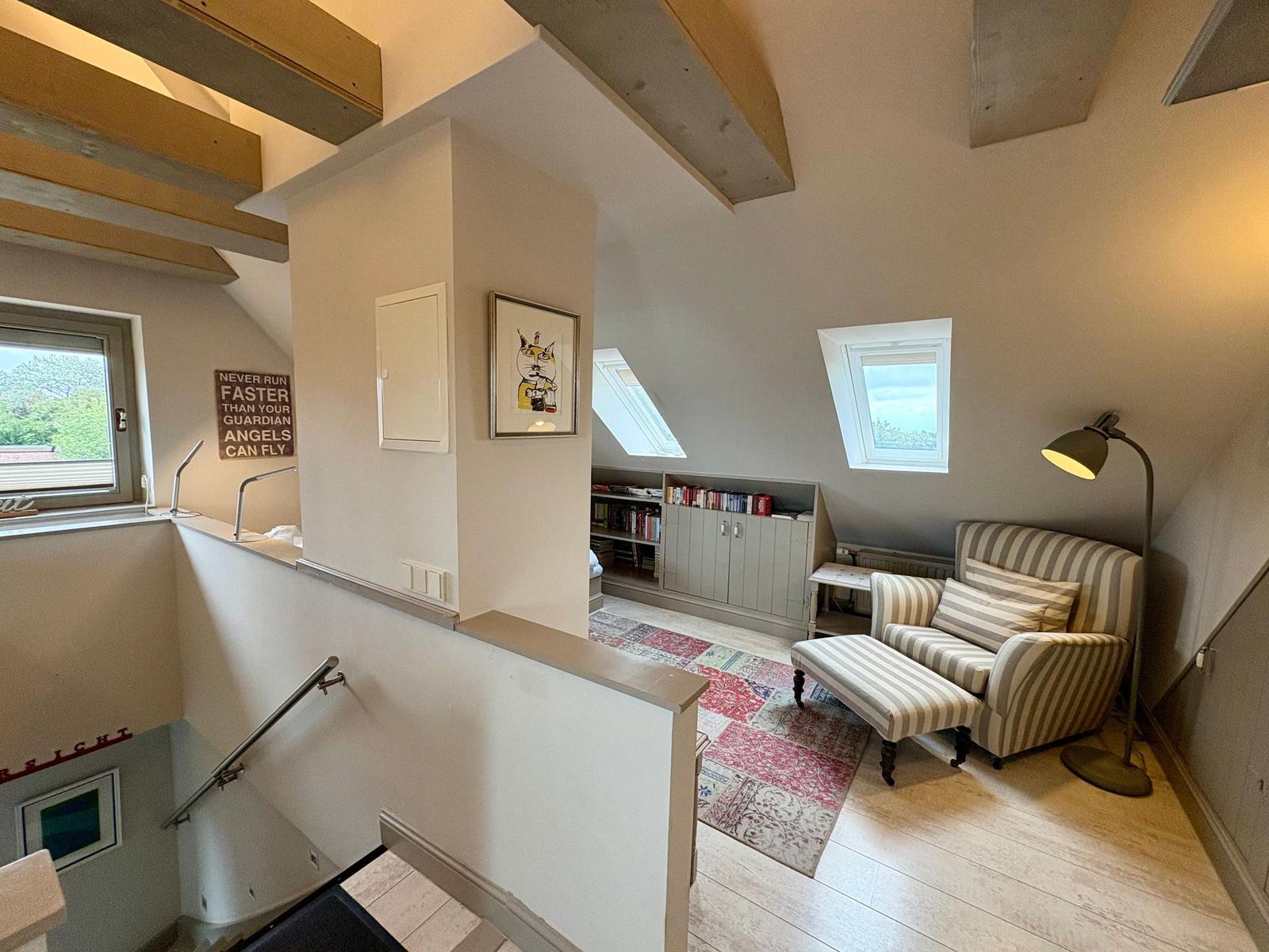 Living area with armchair, windows, and bookcase under wooden beams.