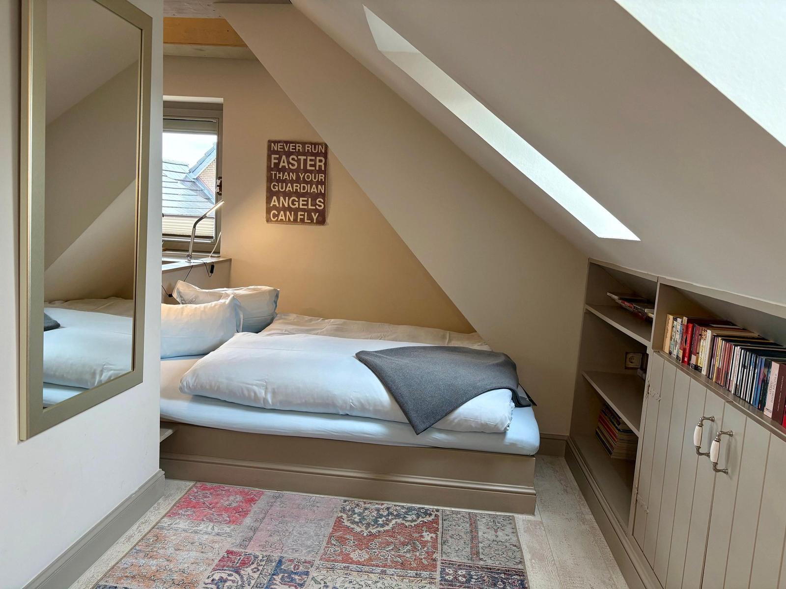 Bedroom with sloped ceiling, bed, mirror, and bookcase under eaves.