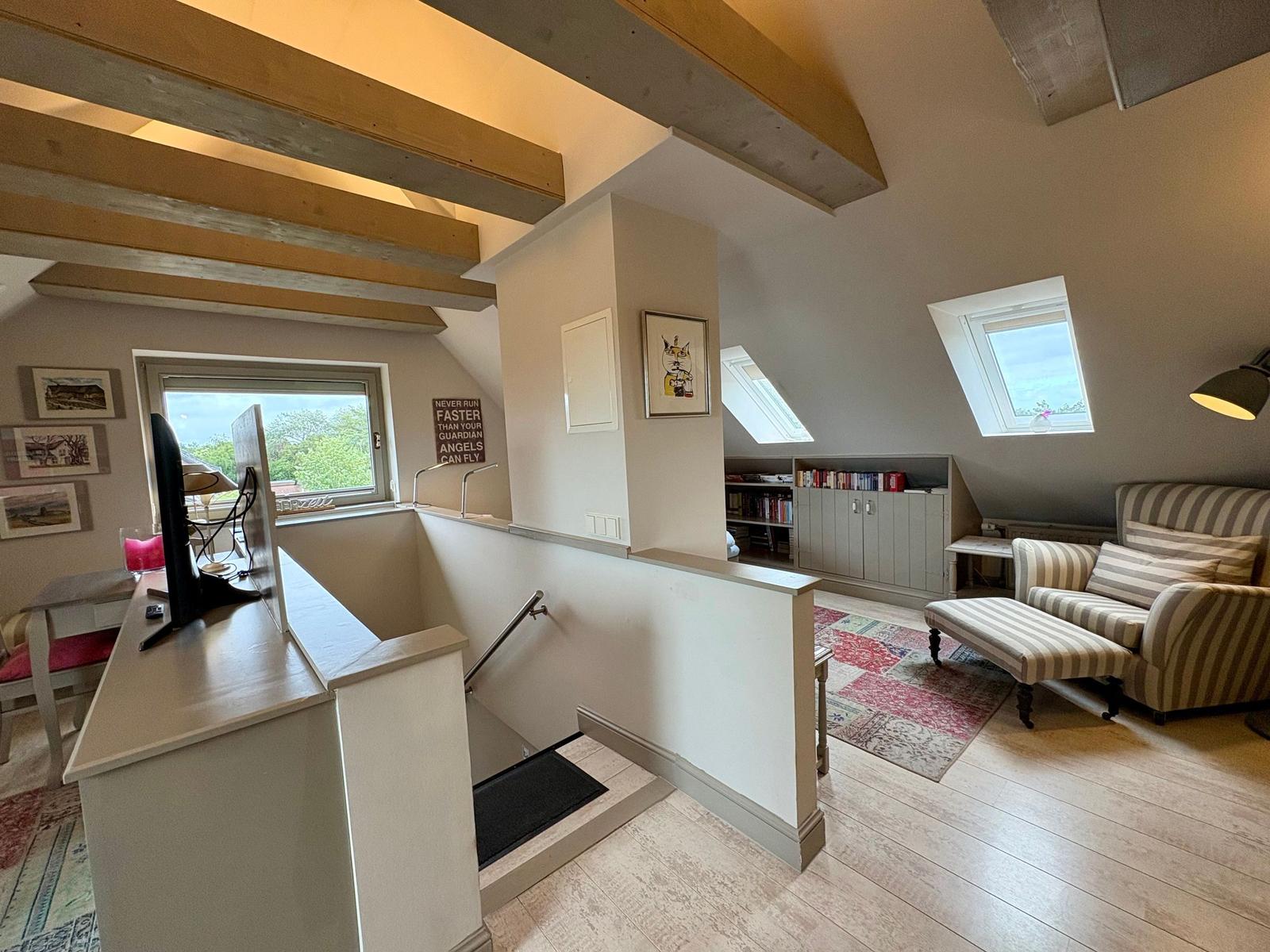 Living area with staircase, armchair, and skylights. Wooden beams and bookcase visible.