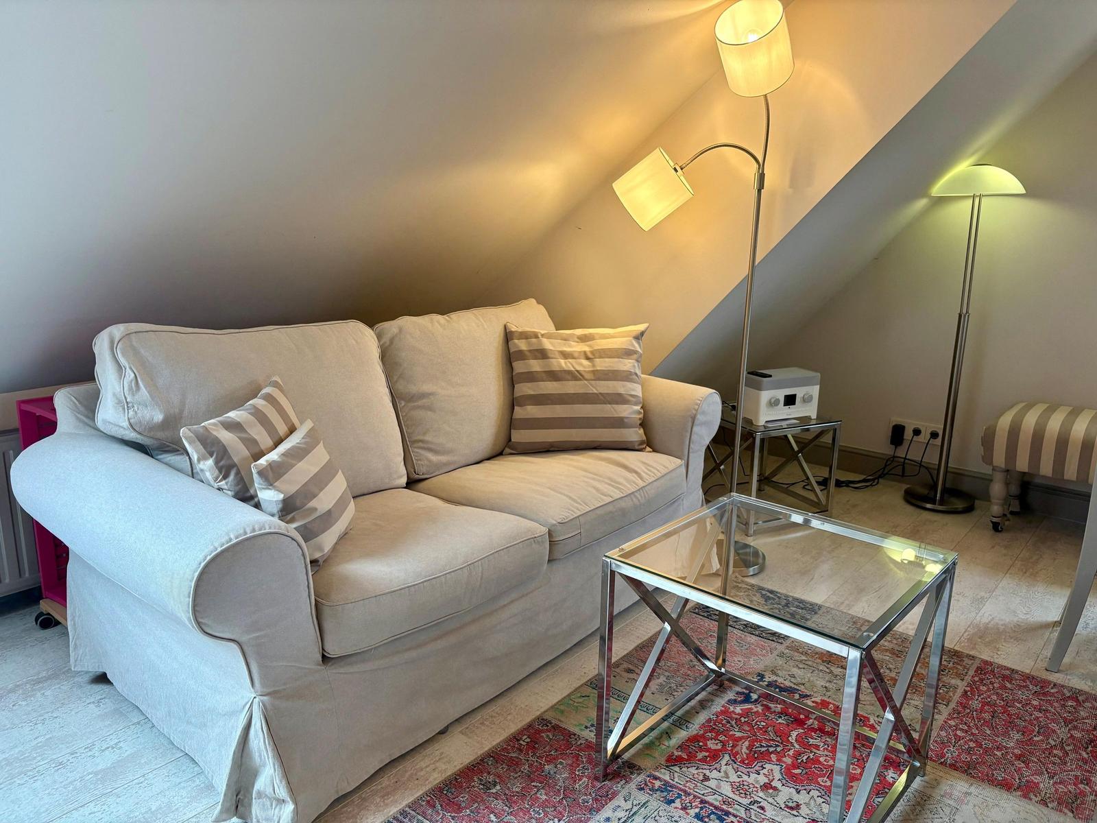 A sofa with cushions, a glass table, and floor lamps in a room with a sloped ceiling.