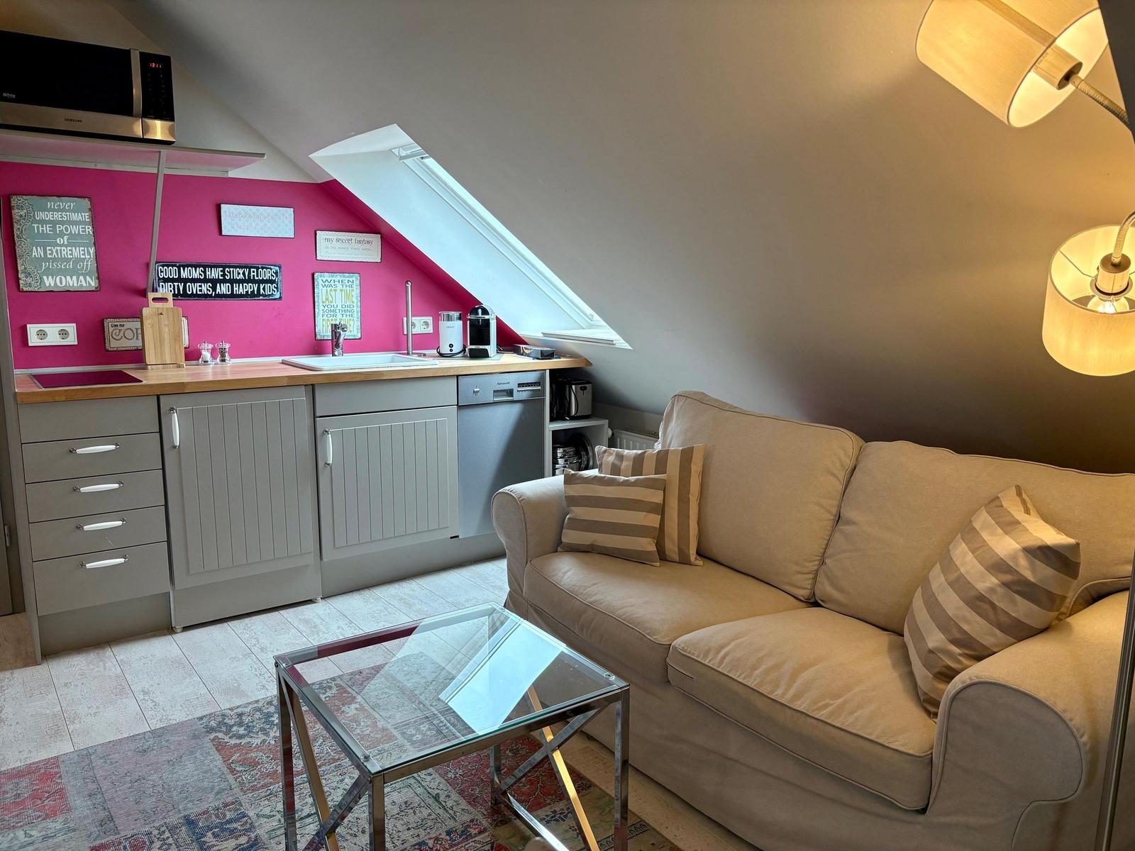 Living area with sofa, glass table, and small kitchen under sloped ceiling.