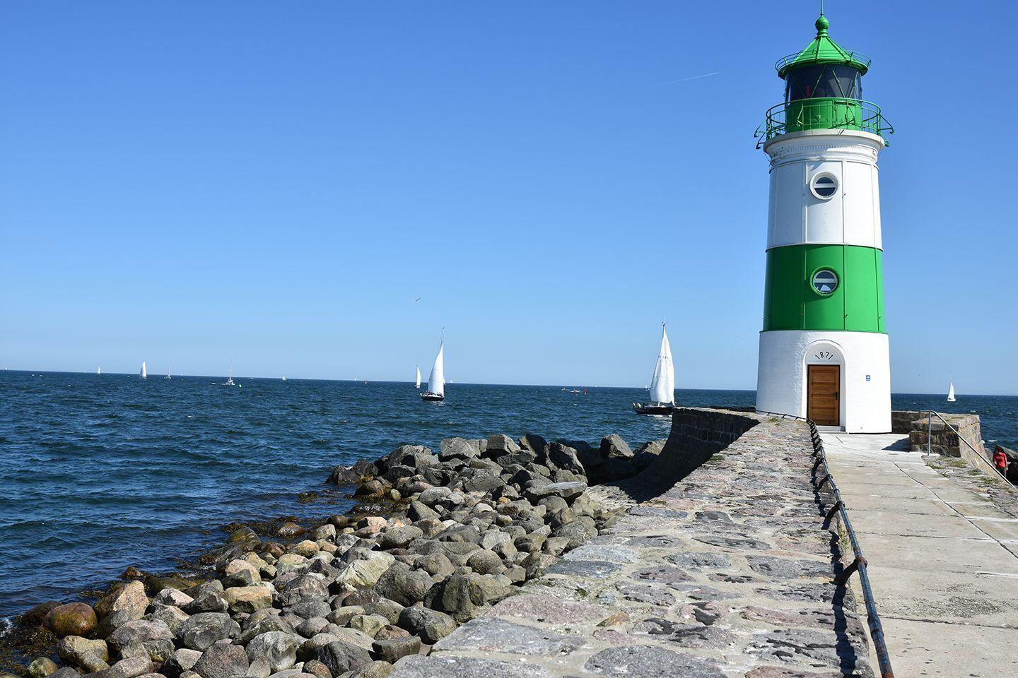 Weiß-grüner Leuchtturm am Steg mit Segelbooten im Hafen.