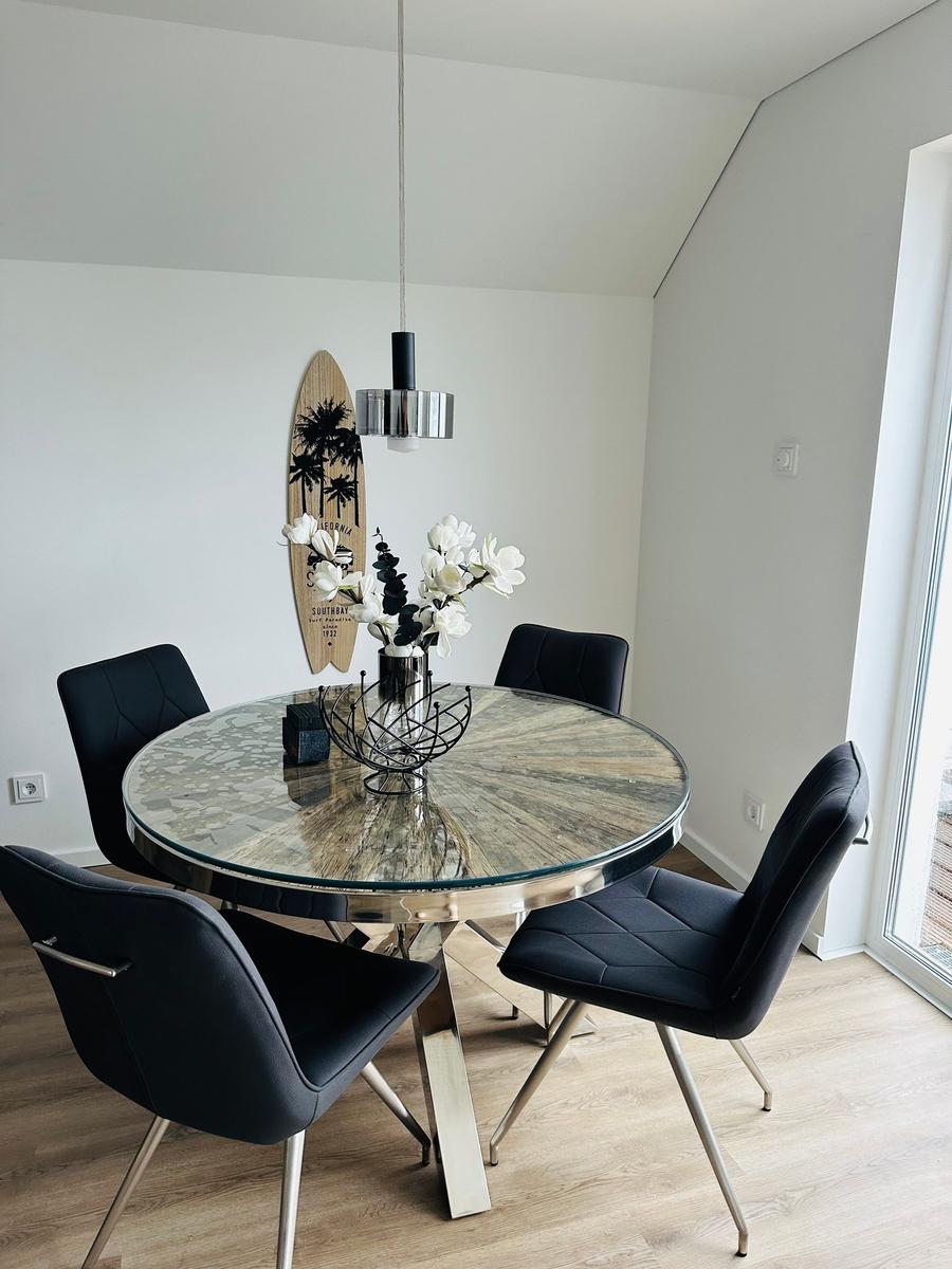 Runder Tisch mit Glasplatte und schwarzen Stühlen. Dekoration mit Blumen und Surfbrett an der Wand.