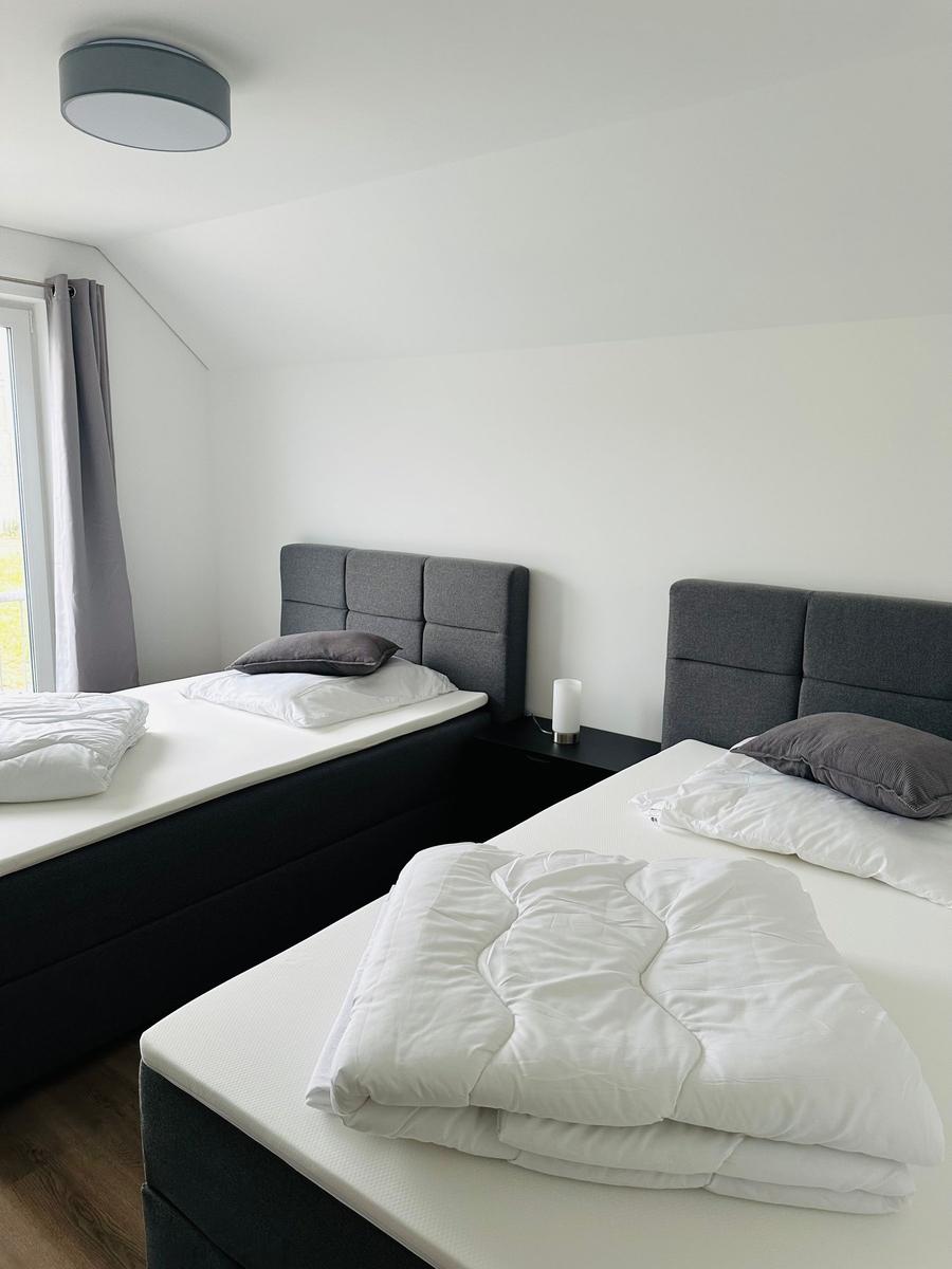 Zwei Betten mit weißen Laken und grauen Kopfkissen. Schwarzer Nachttisch und Fenster mit grauen Vorhängen.