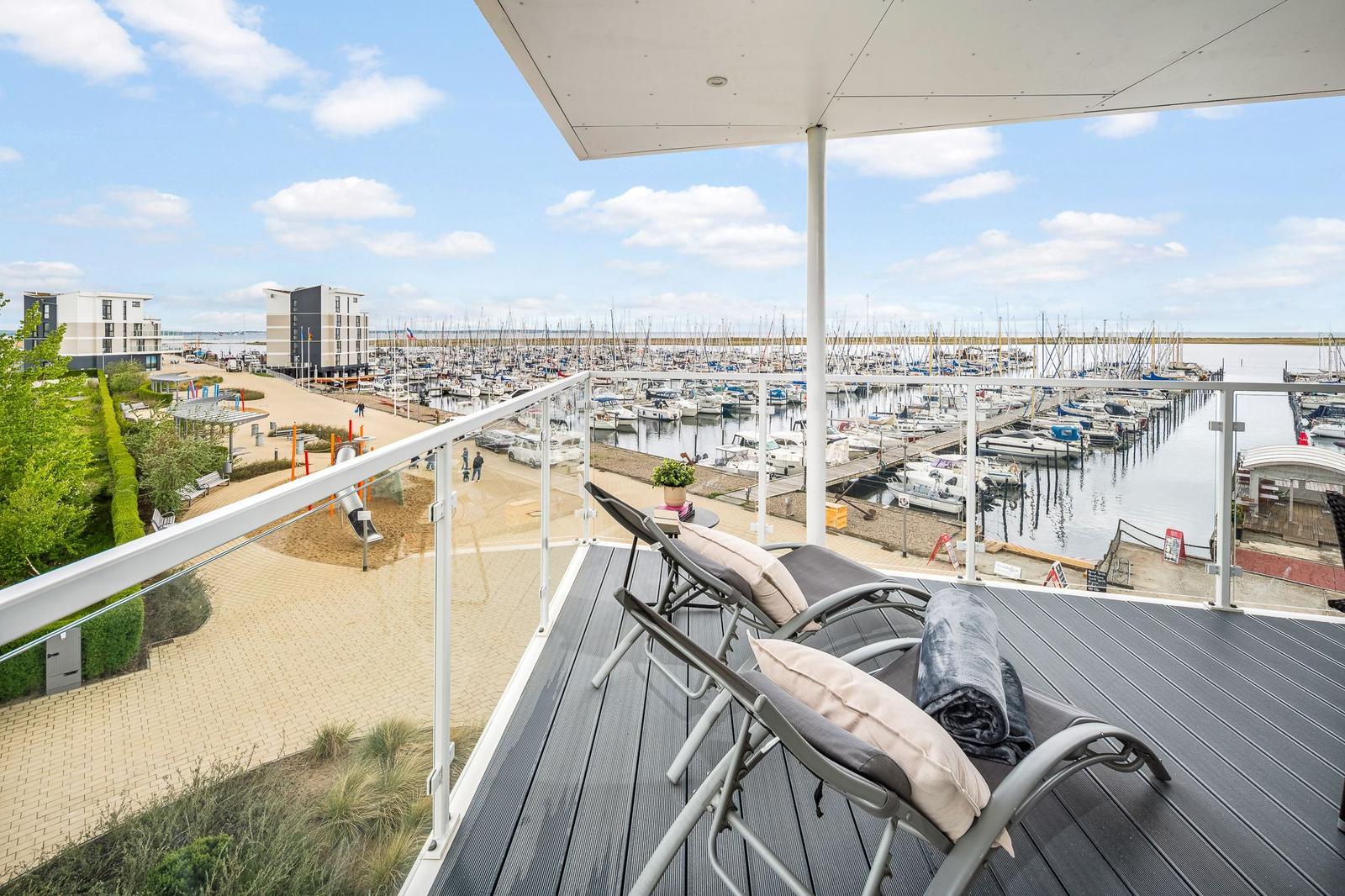 Terrasse mit Liegestühlen und Blick auf Hafen mit Booten und Strandbereich.