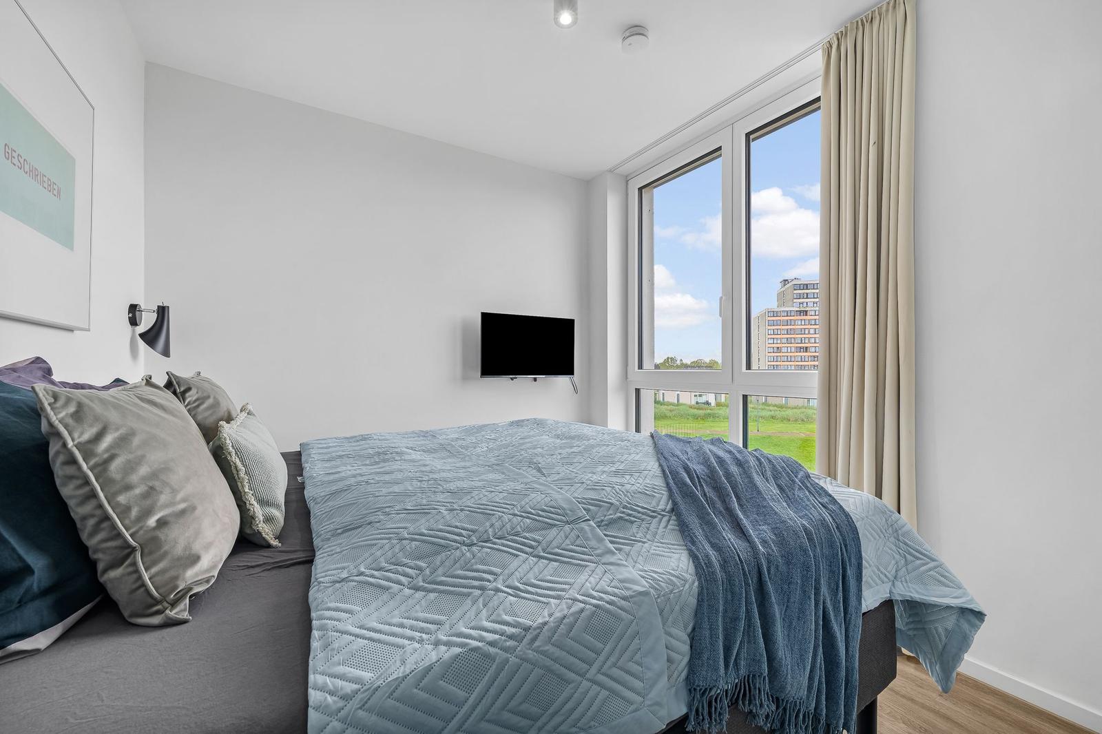 Schlafzimmer mit Bett, TV und großem Fenster mit Blick auf Grünfläche und Gebäude.