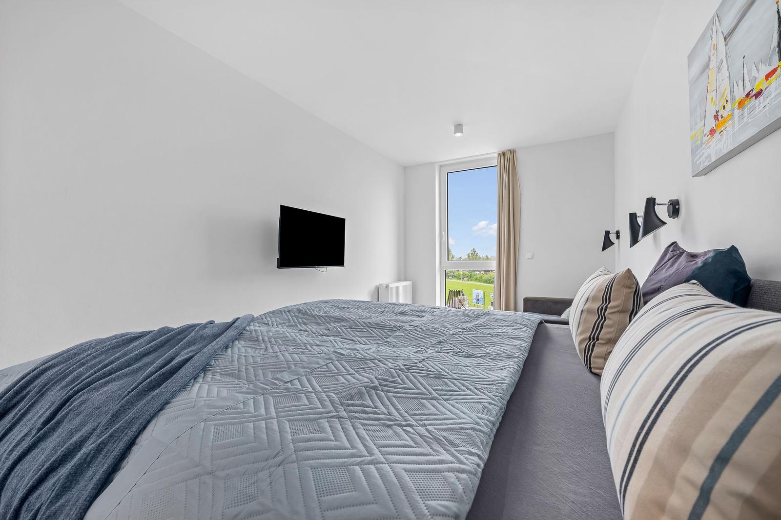 Schlafzimmer mit Bett, Fernseher und Fenster mit Blick auf Grünfläche.