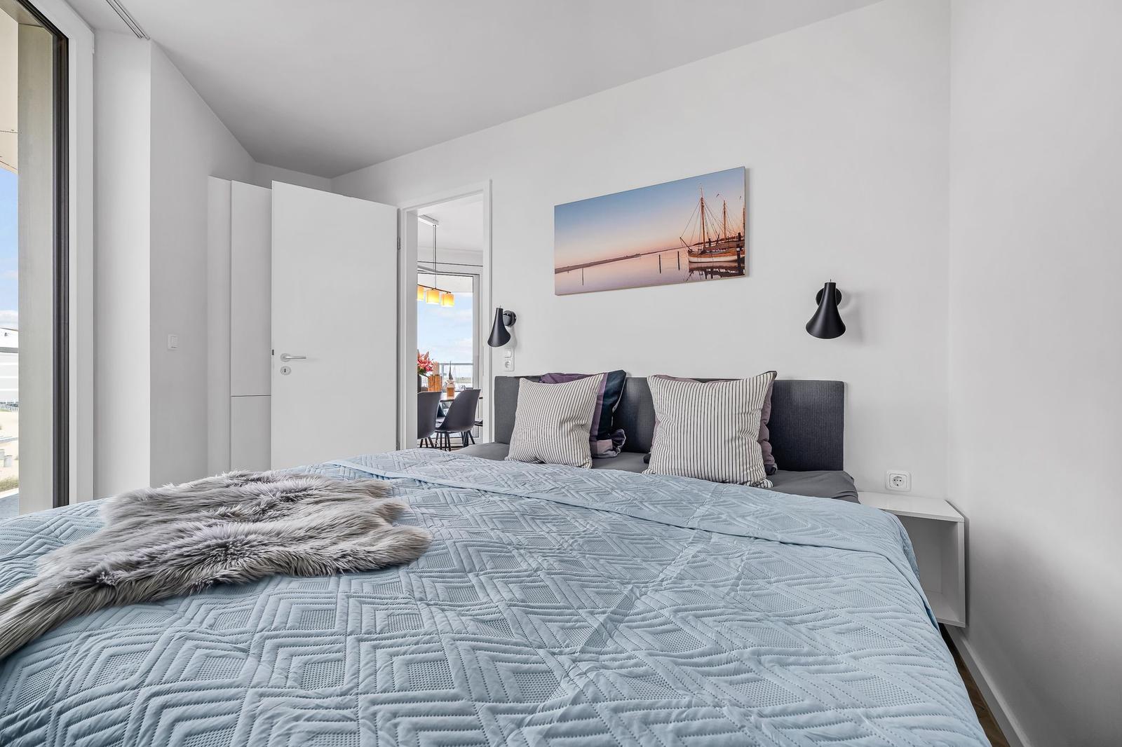 Schlafzimmer mit Bett, Wandbild und Nachttisch. Tür führt in Wohnbereich.