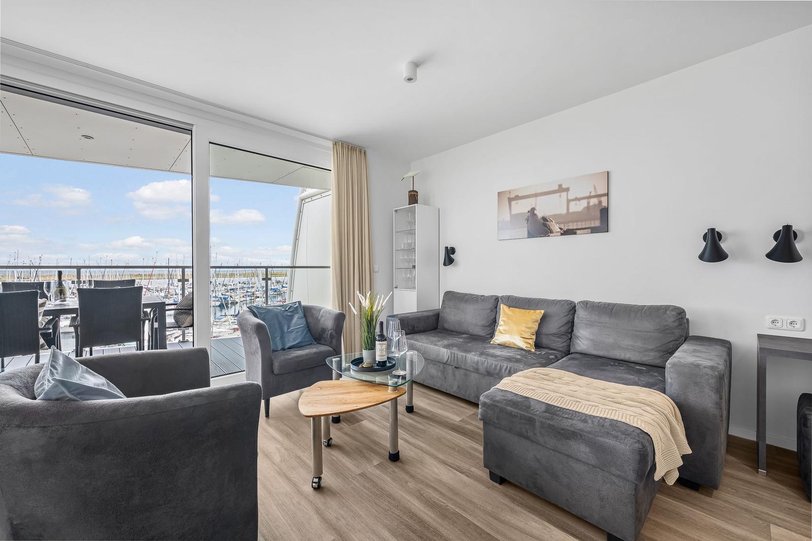 Wohnzimmer mit grauem Sofa, Glas-Tisch und Balkonblick auf Hafen.