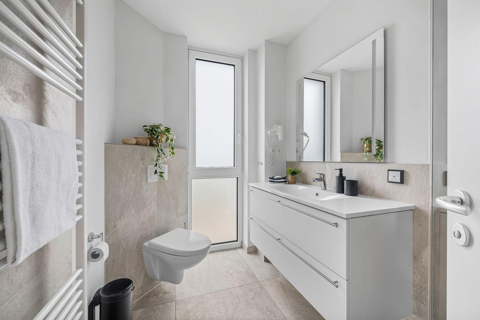 Badezimmer mit Waschtisch, Spiegel, Toilette und Heizung. Fenster sorgt für Tageslicht.