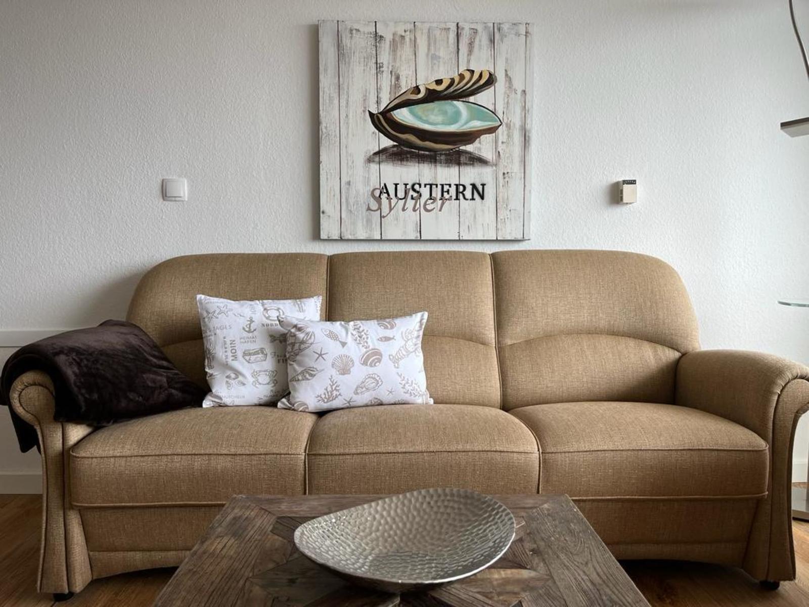 Beige Sofa mit Kissen und Wandbild mit Muschel. Holztisch mit Metallschale.