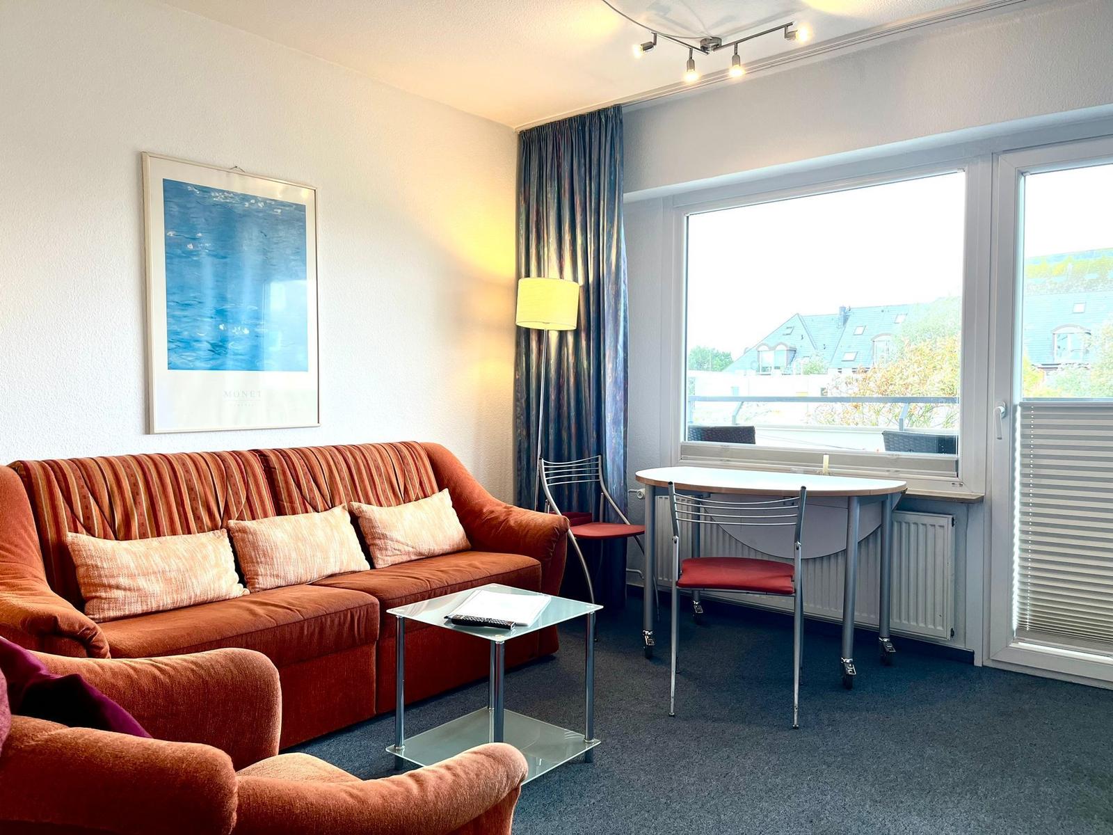 Wohnzimmer mit Sofa, Tisch und Stühlen. Fenster mit Blick auf Balkon und Gebäude.