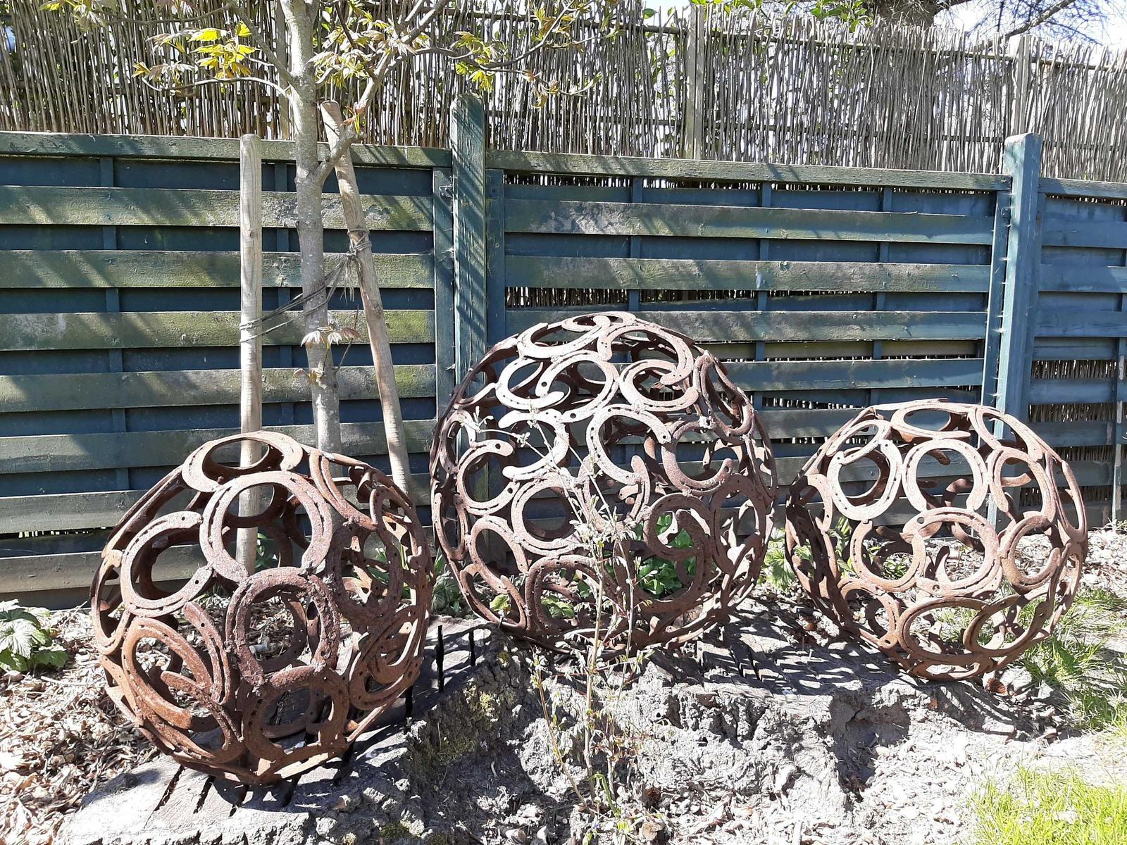 Drei runde Skulpturen aus Hufeisen stehen im Garten vor einem Holzzaun.